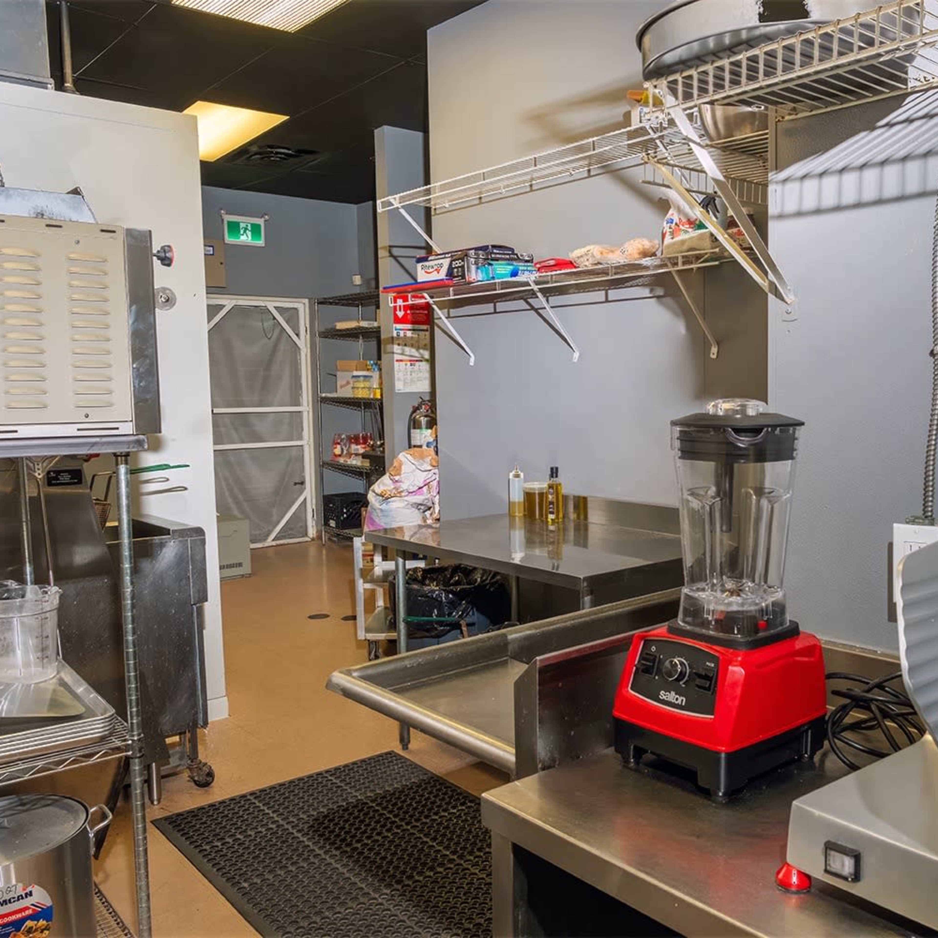 A commercial kitchen setup with stainless steel counters, a blender, a meat slicer, and various kitchen utensils on a wall-mounted rack.