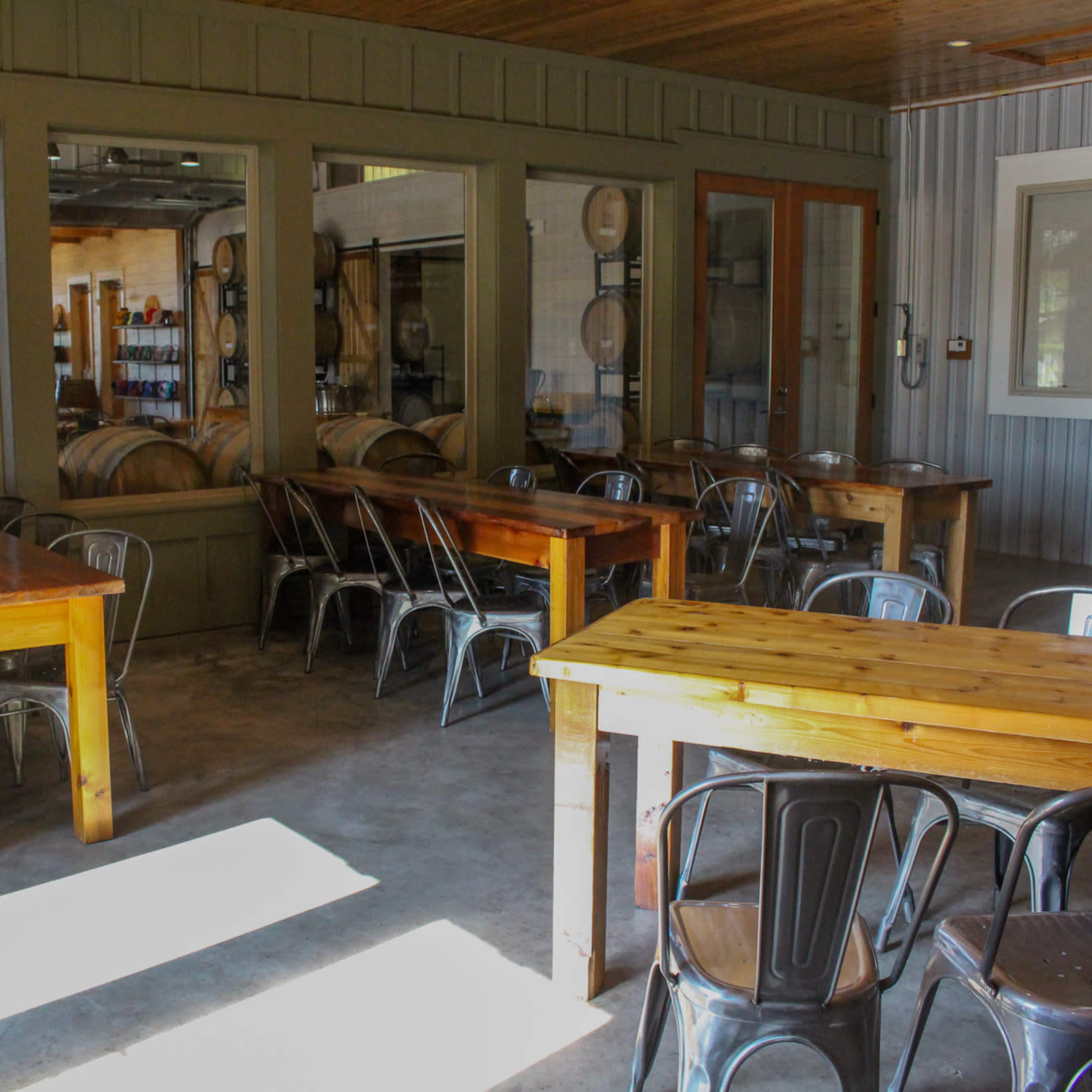 A dining area with wooden tables and metal chairs, alongside a view of storage barrels through large windows.