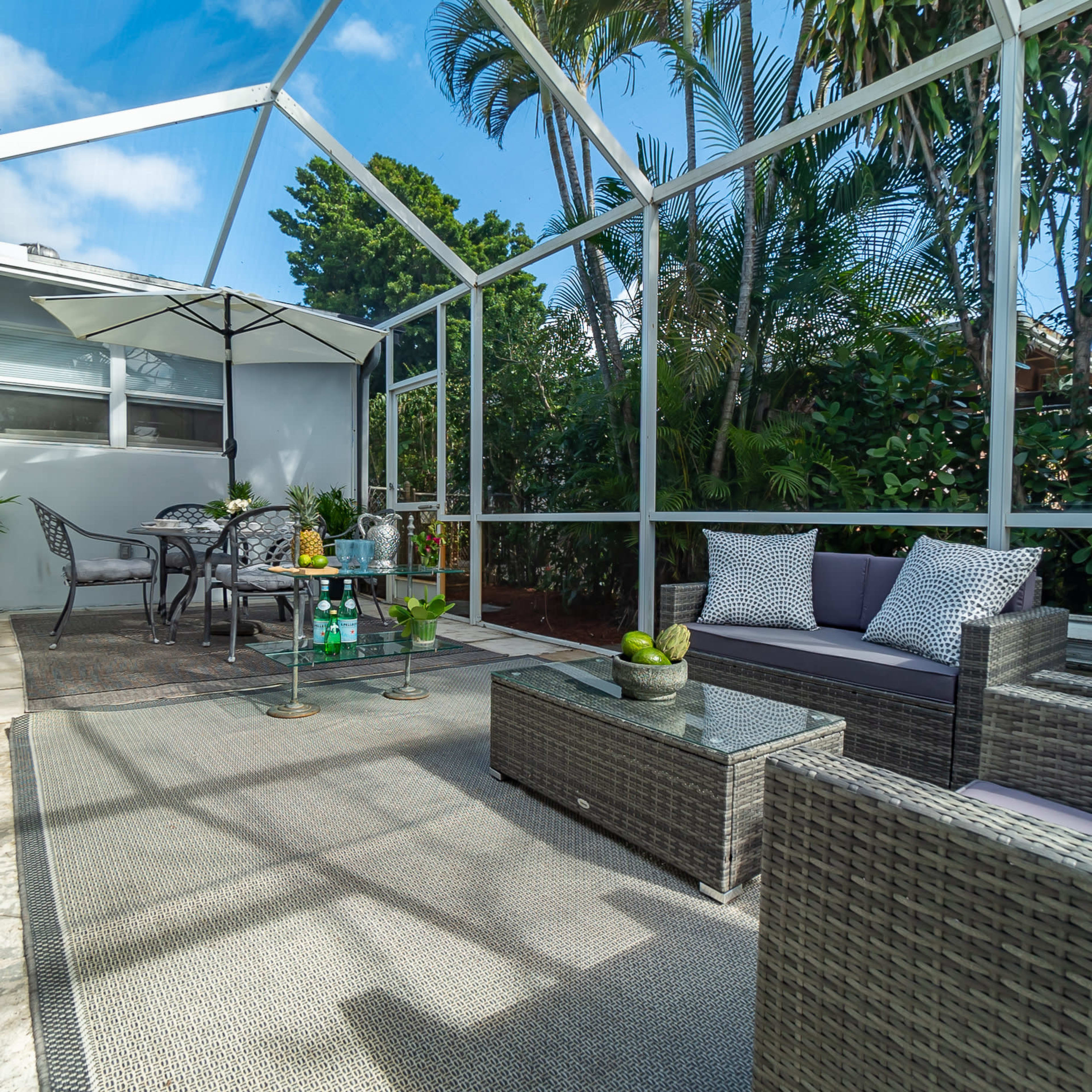 A screened-in patio features a seating area with wicker furniture, a table, and an umbrella, surrounded by greenery.