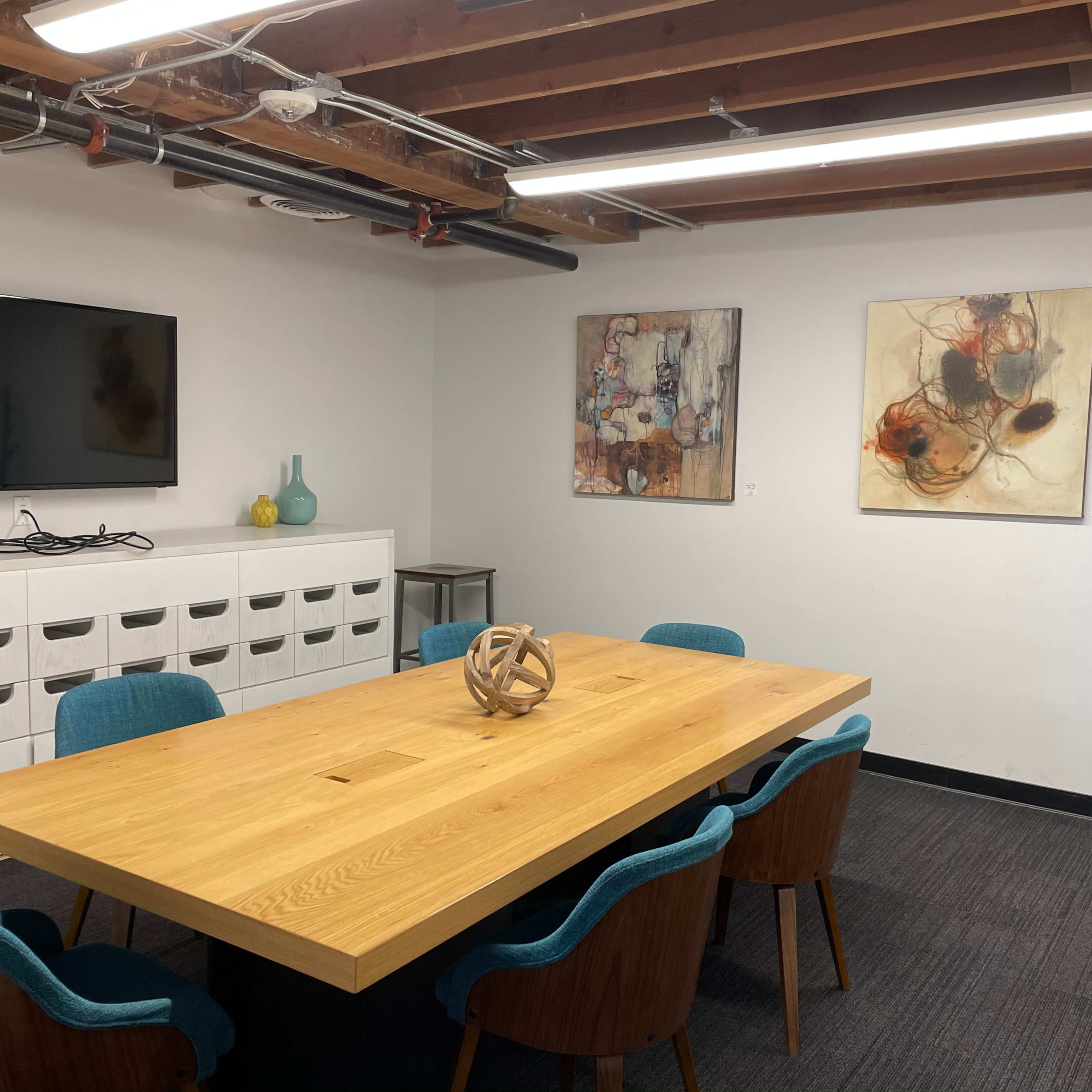 A conference room features a long wooden table surrounded by blue chairs, with a television mounted on the wall and two abstract art pieces hanging nearby.