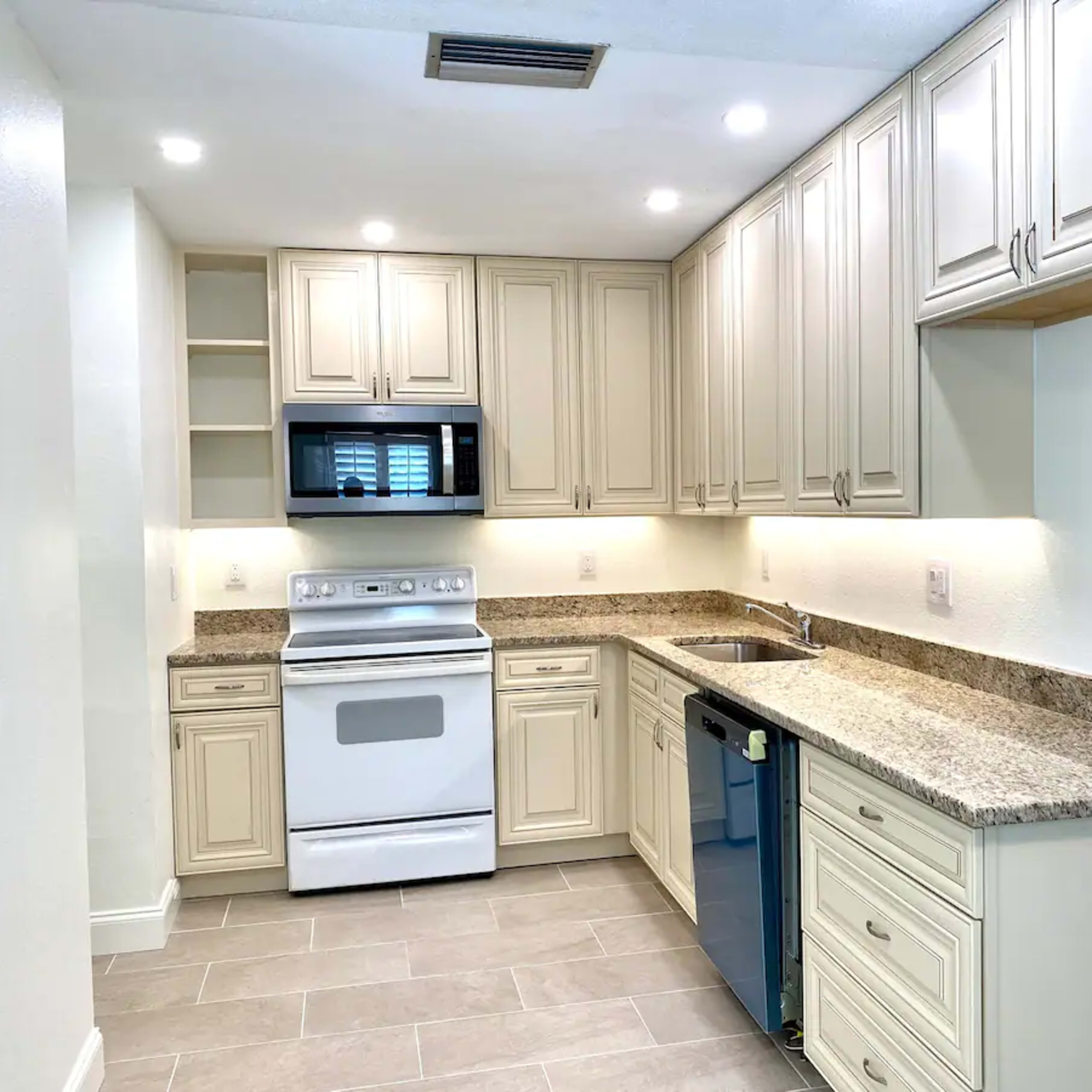 A modern kitchen with light-colored cabinets, granite countertops, a white stove, and a blue dishwasher.