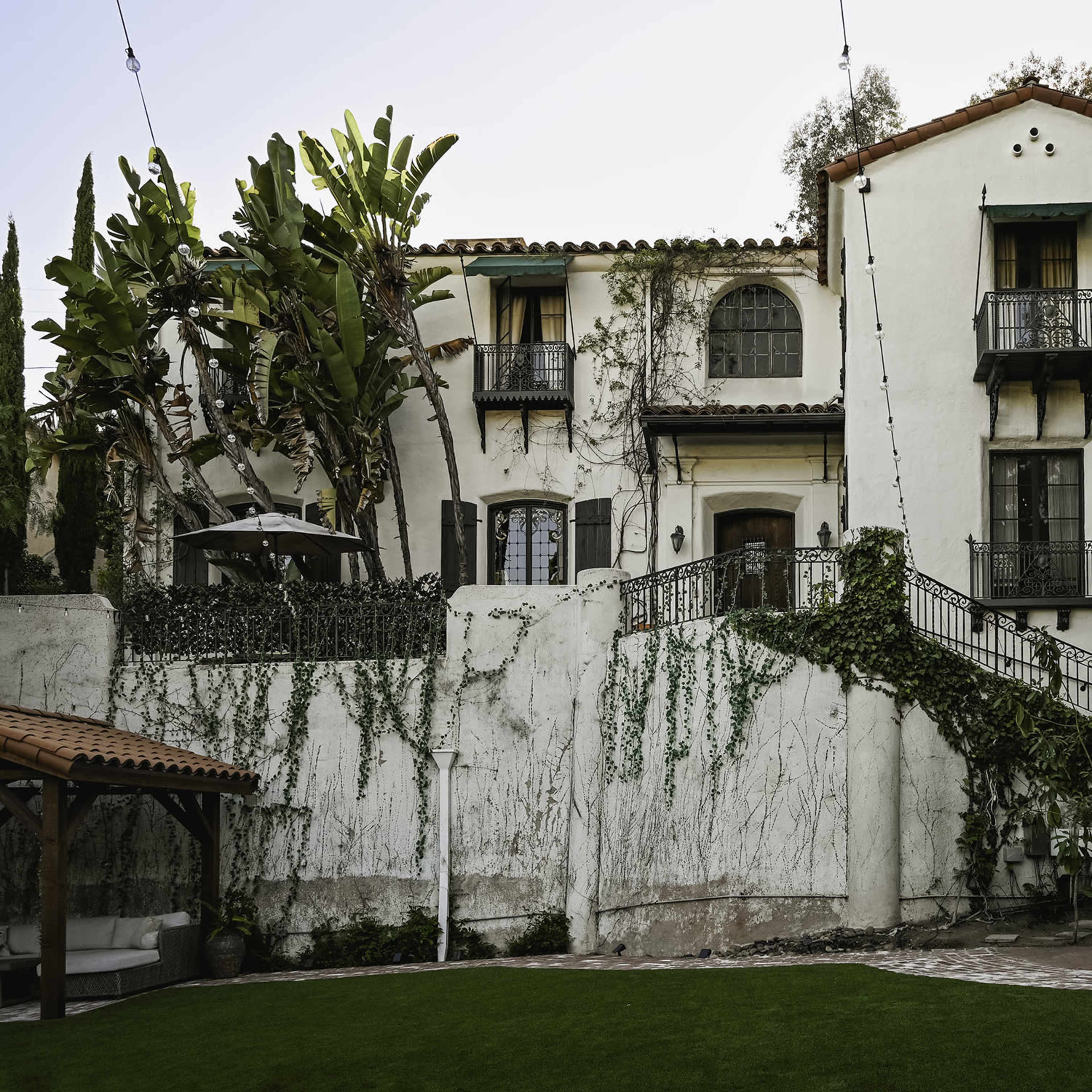 A multi-story Mediterranean-style house with balconies, surrounded by greenery and a landscaped yard.