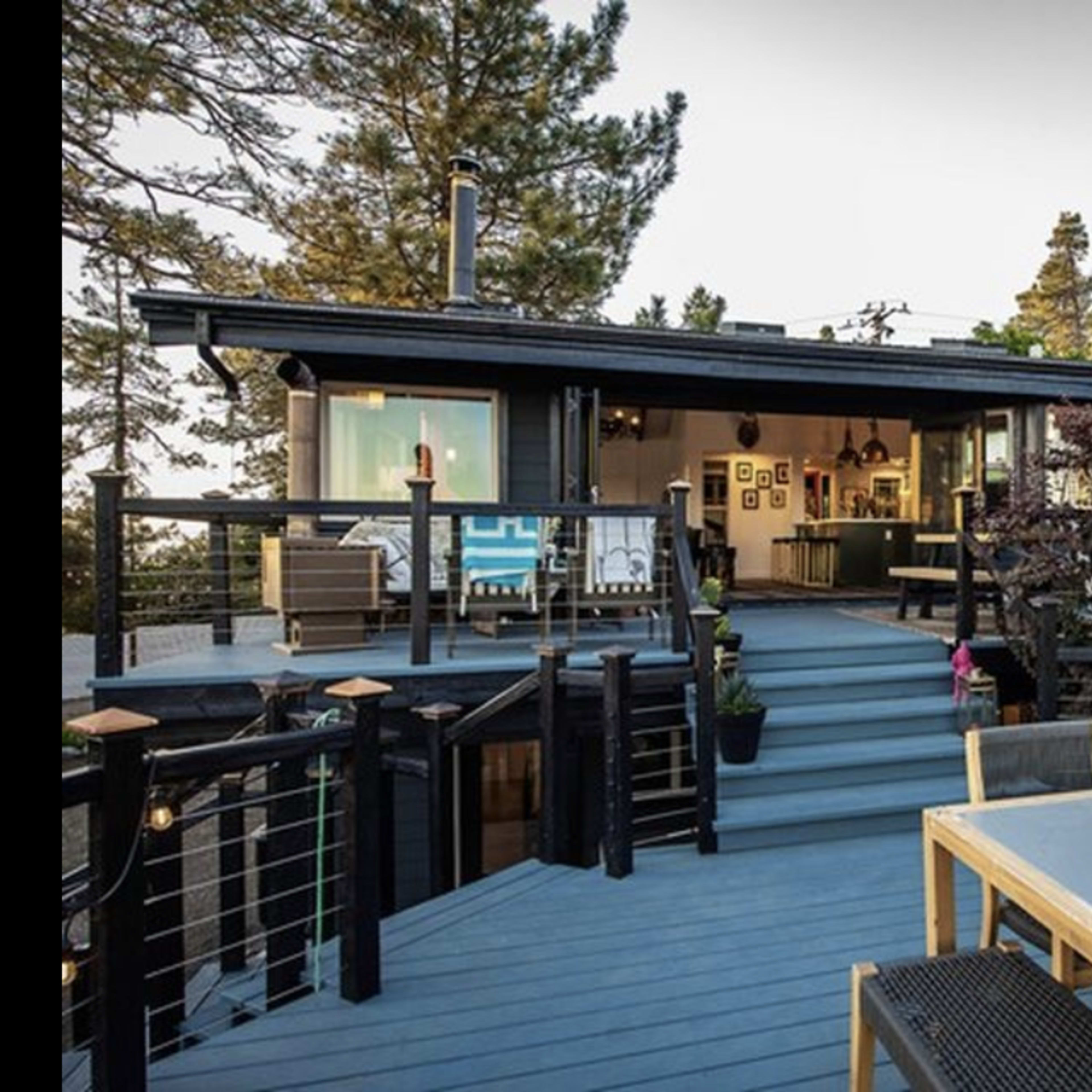 A modern wooden deck with a dining table, leading to a contemporary house surrounded by trees.