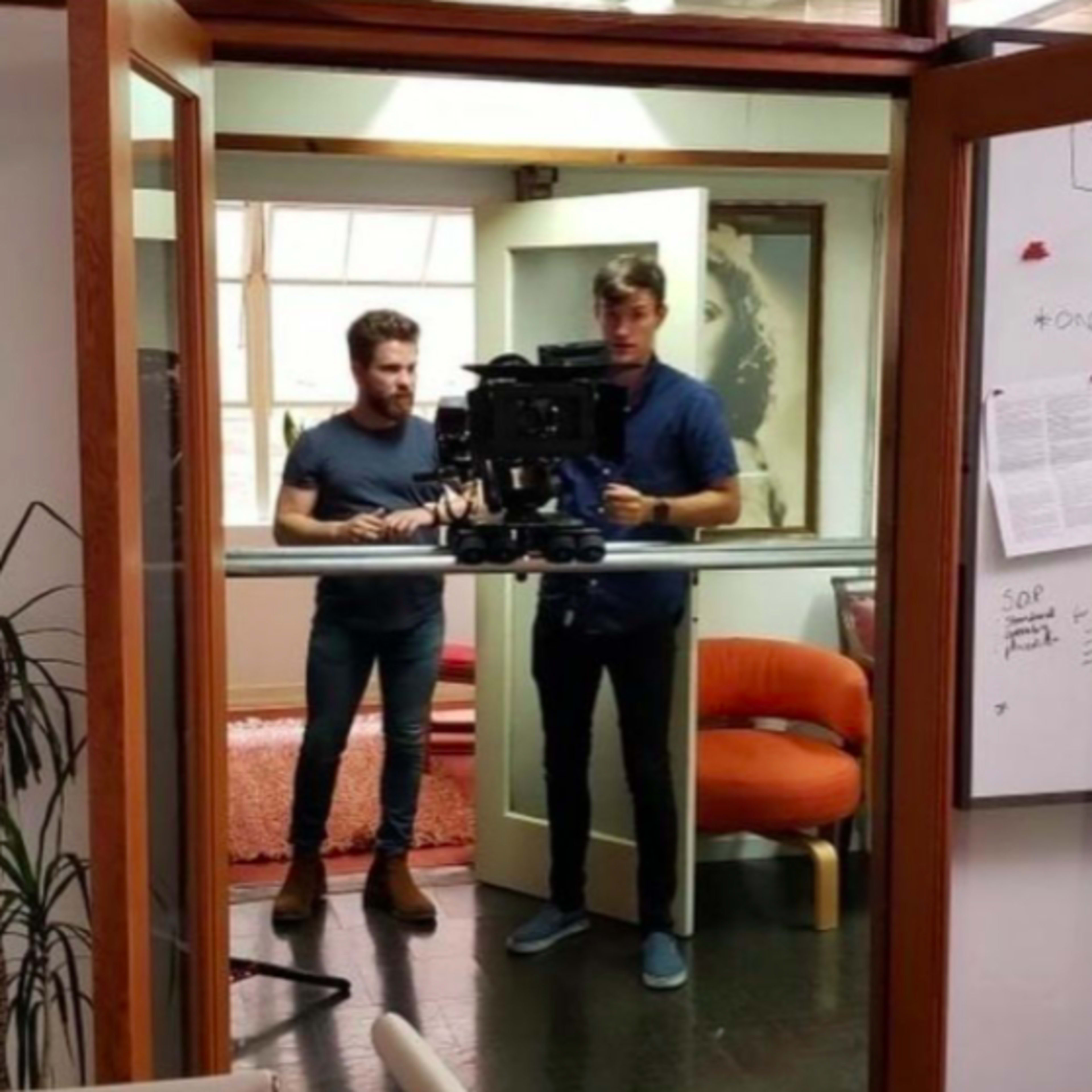 Two men are filming in an office setting, positioned in front of a camera mounted on a tripod.