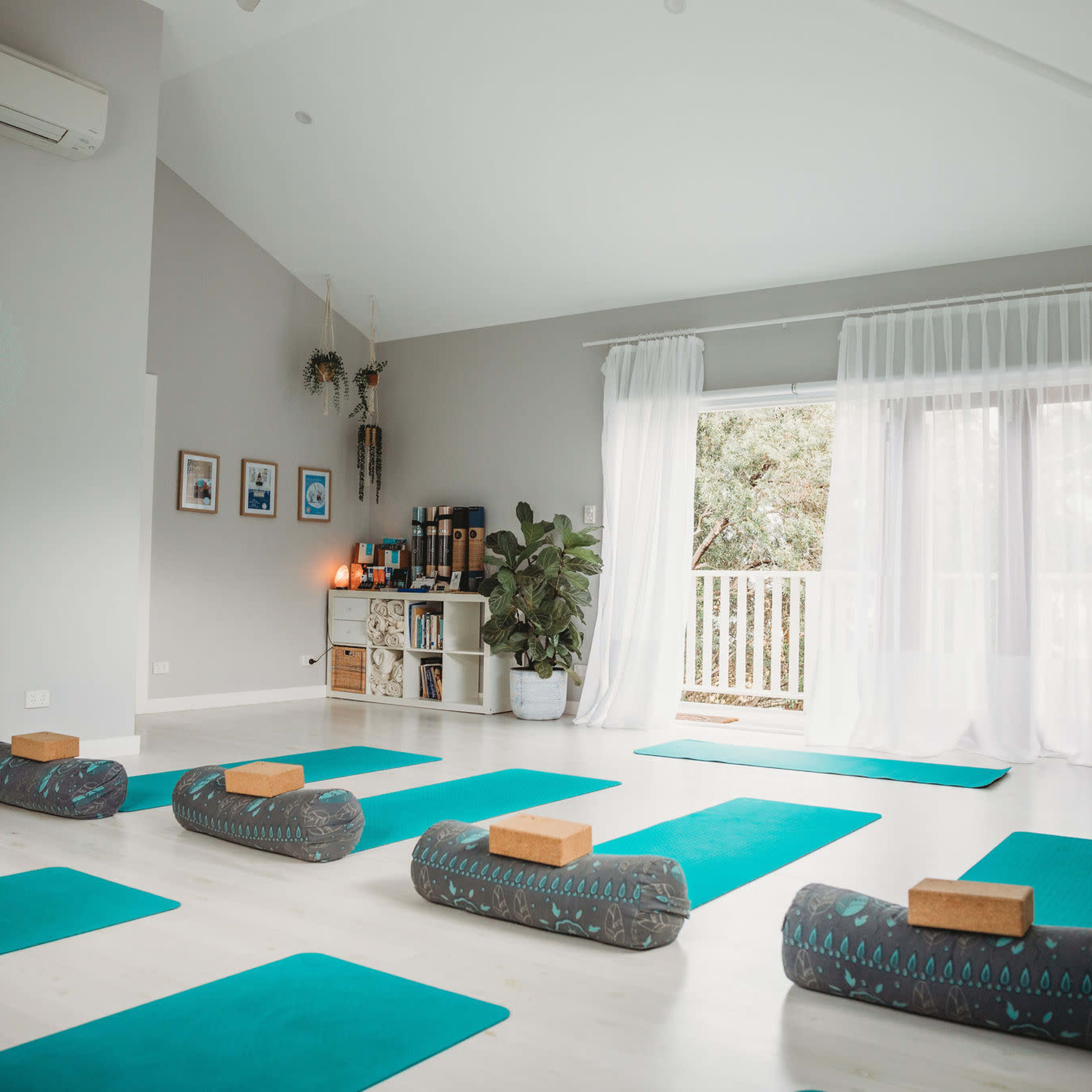 A spacious yoga studio is set up with green mats and bolsters facing a large window, with plants and decor lining the walls.