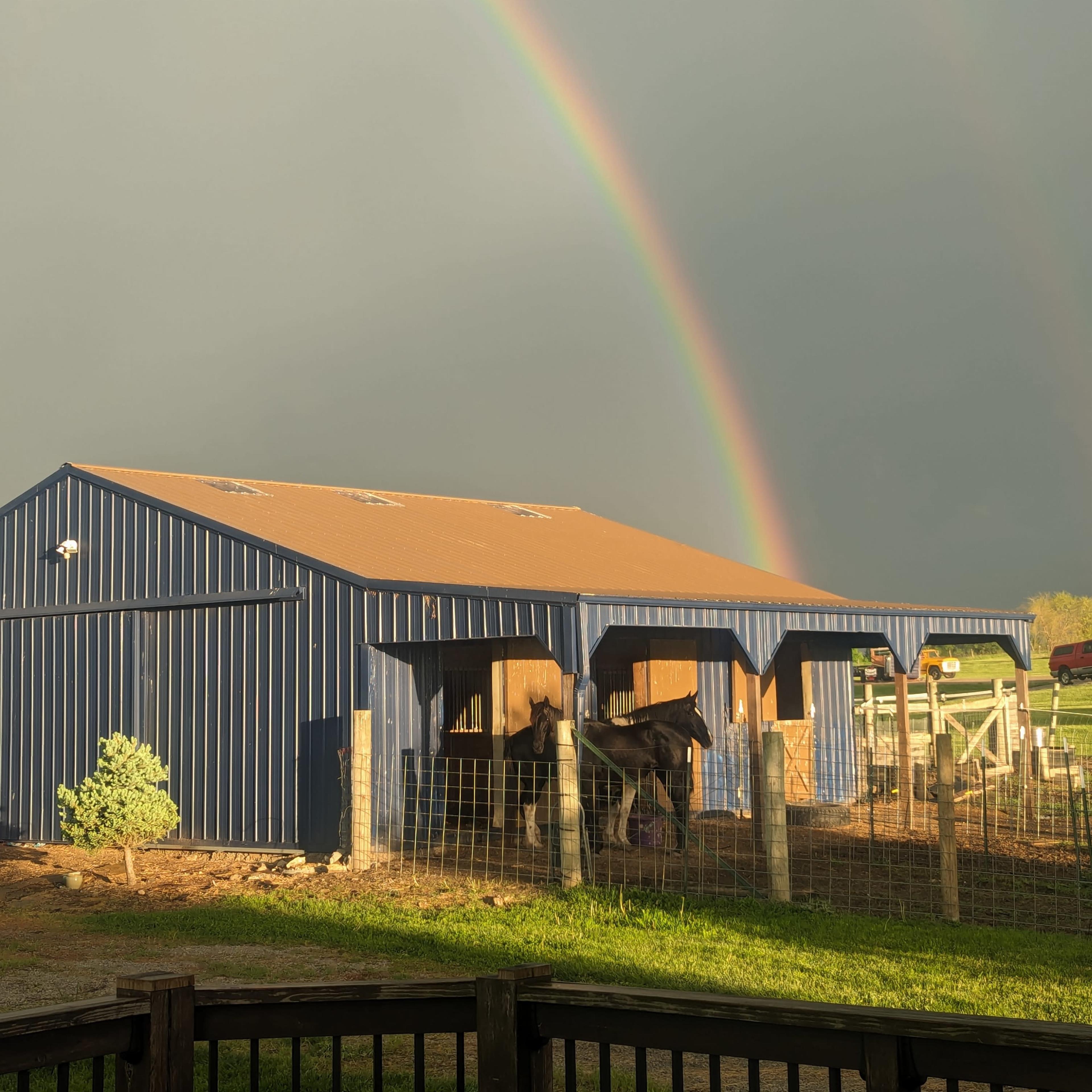 A blue barn stands under a double rainbow, with a horse visible in a nearby pen.