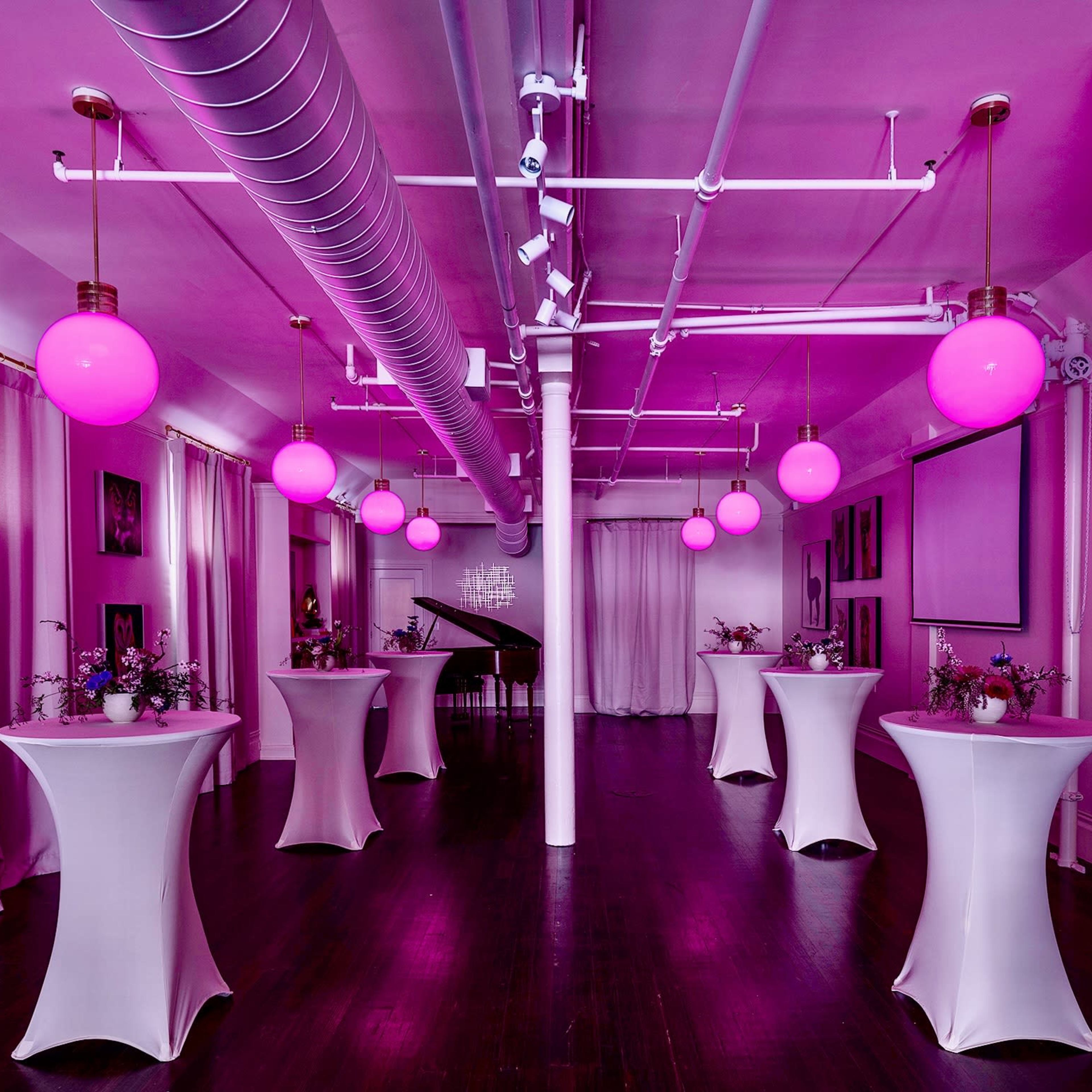 A spacious event room features several tall tables adorned with flowers and illuminated by pink globe pendant lights.