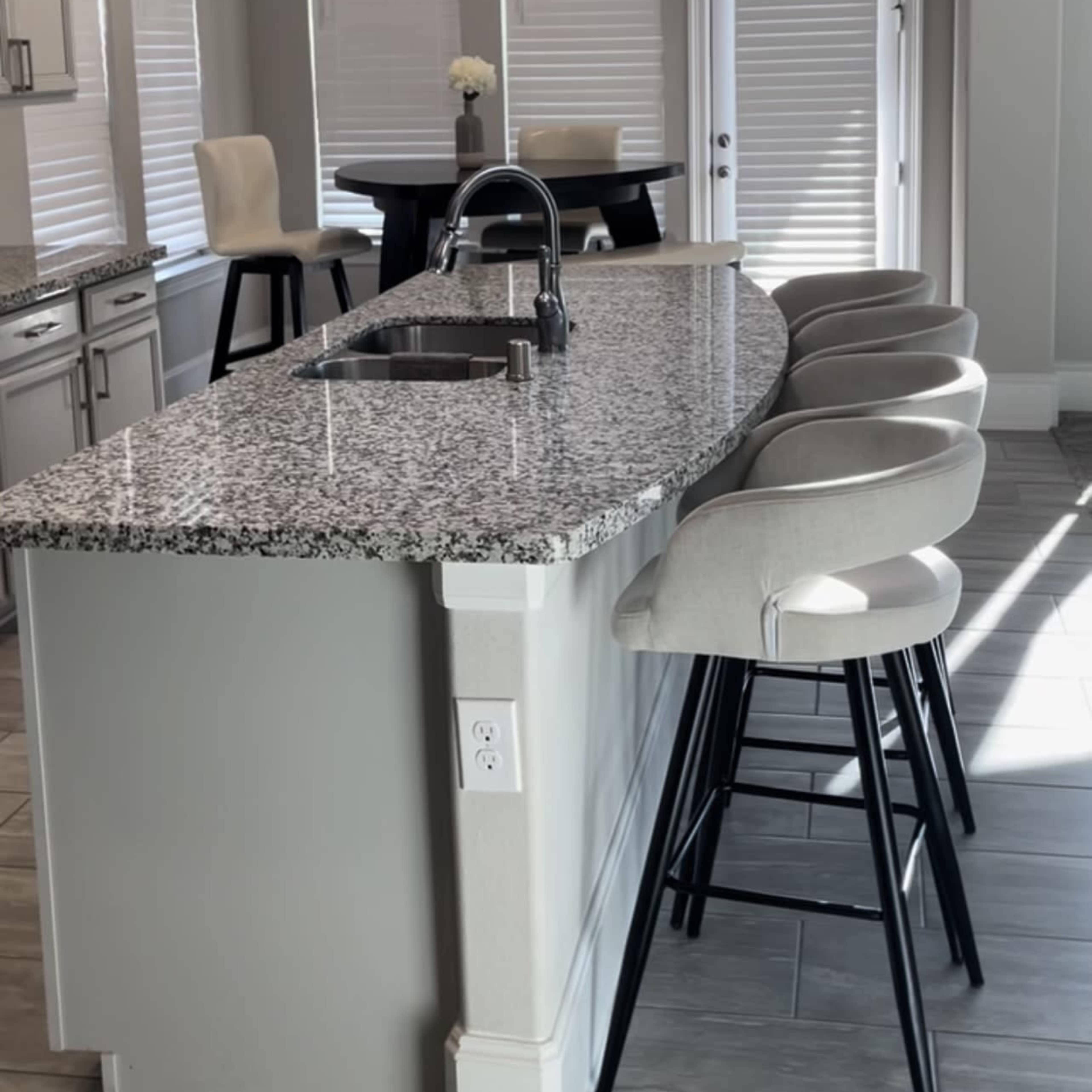 A modern kitchen features a curved granite countertop with four bar stools, pendant lights above, and large windows allowing natural light.