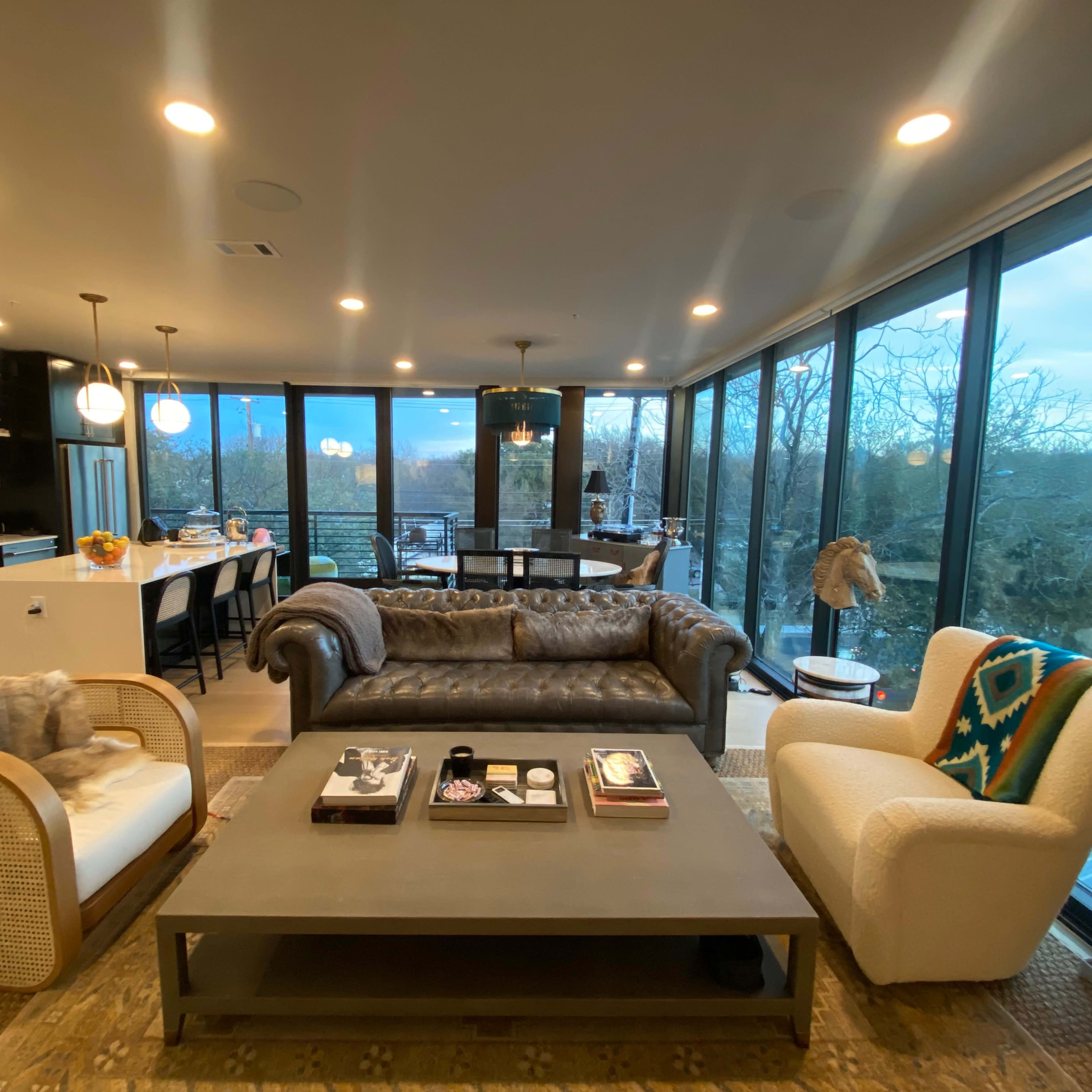 A modern living area with large windows, featuring a brown leather sofa, two armchairs, and a coffee table, all set in an open-concept layout that connects to a kitchen.