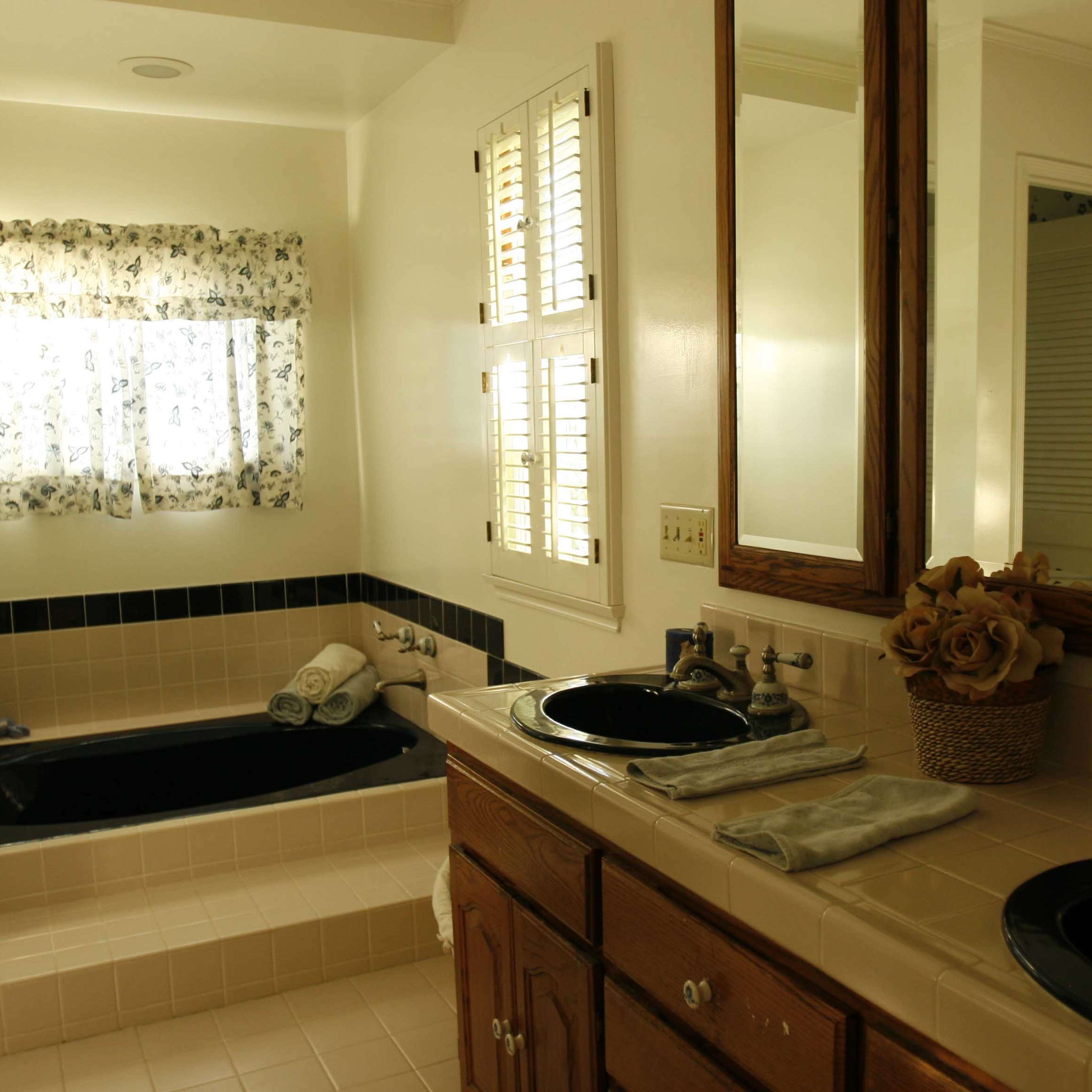 A spacious bathroom with a dark bathtub, dual sinks, wooden cabinets, and a window covered with floral curtains.