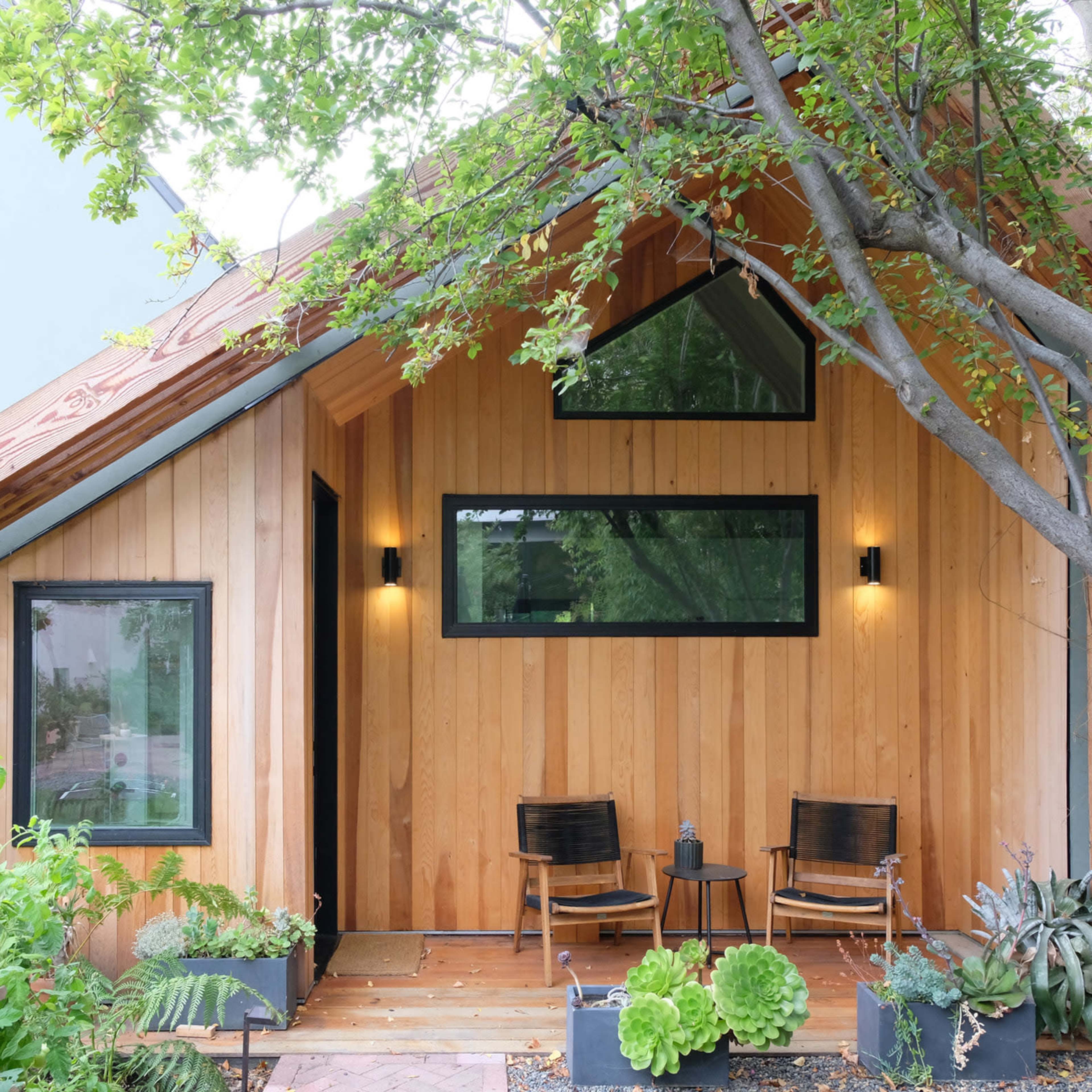 A wooden exterior of a modern house with two chairs on the porch, surrounded by plants and trees.