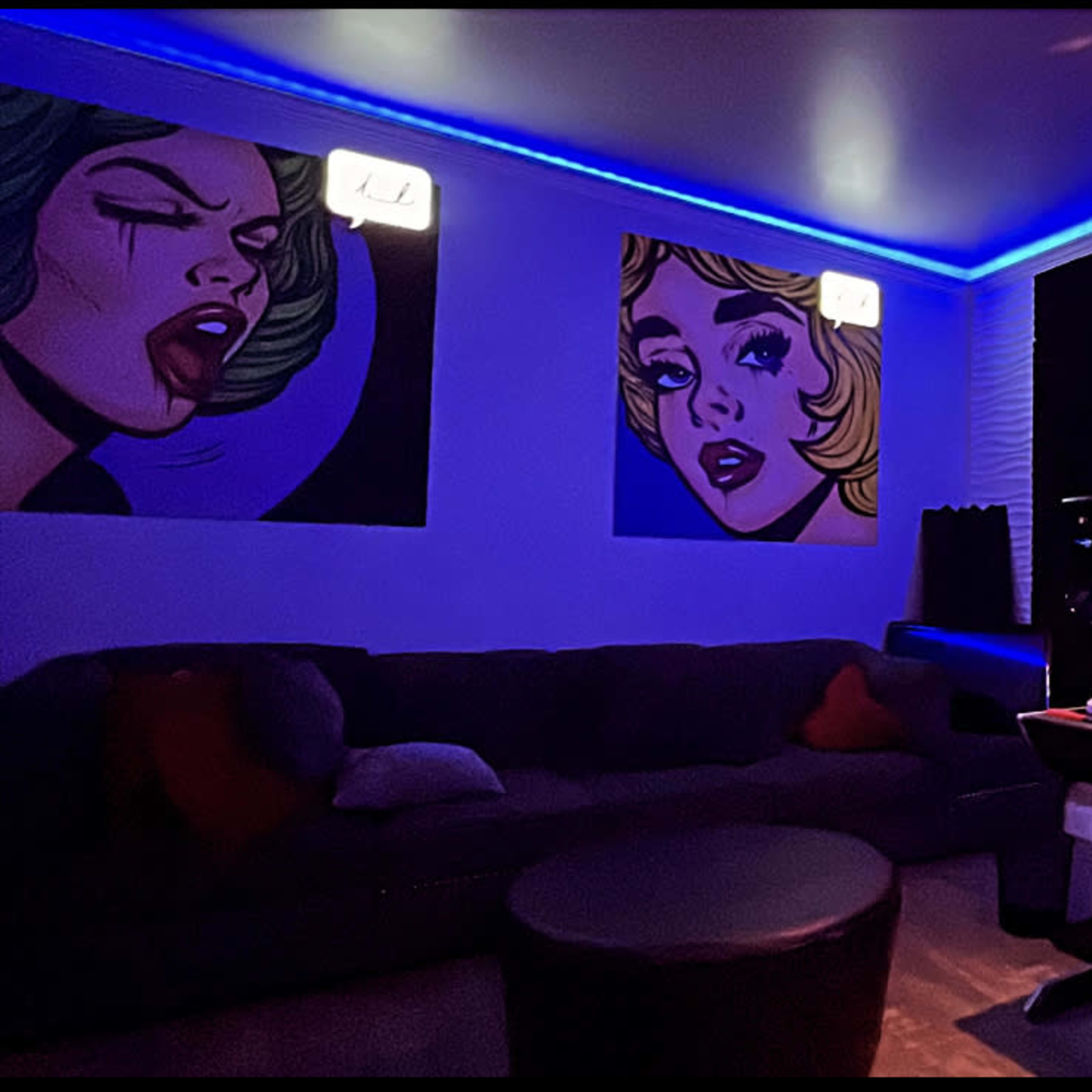 A modern music studio with colorful wall art, a dark couch, and a desk equipped with audio equipment under blue LED lighting.