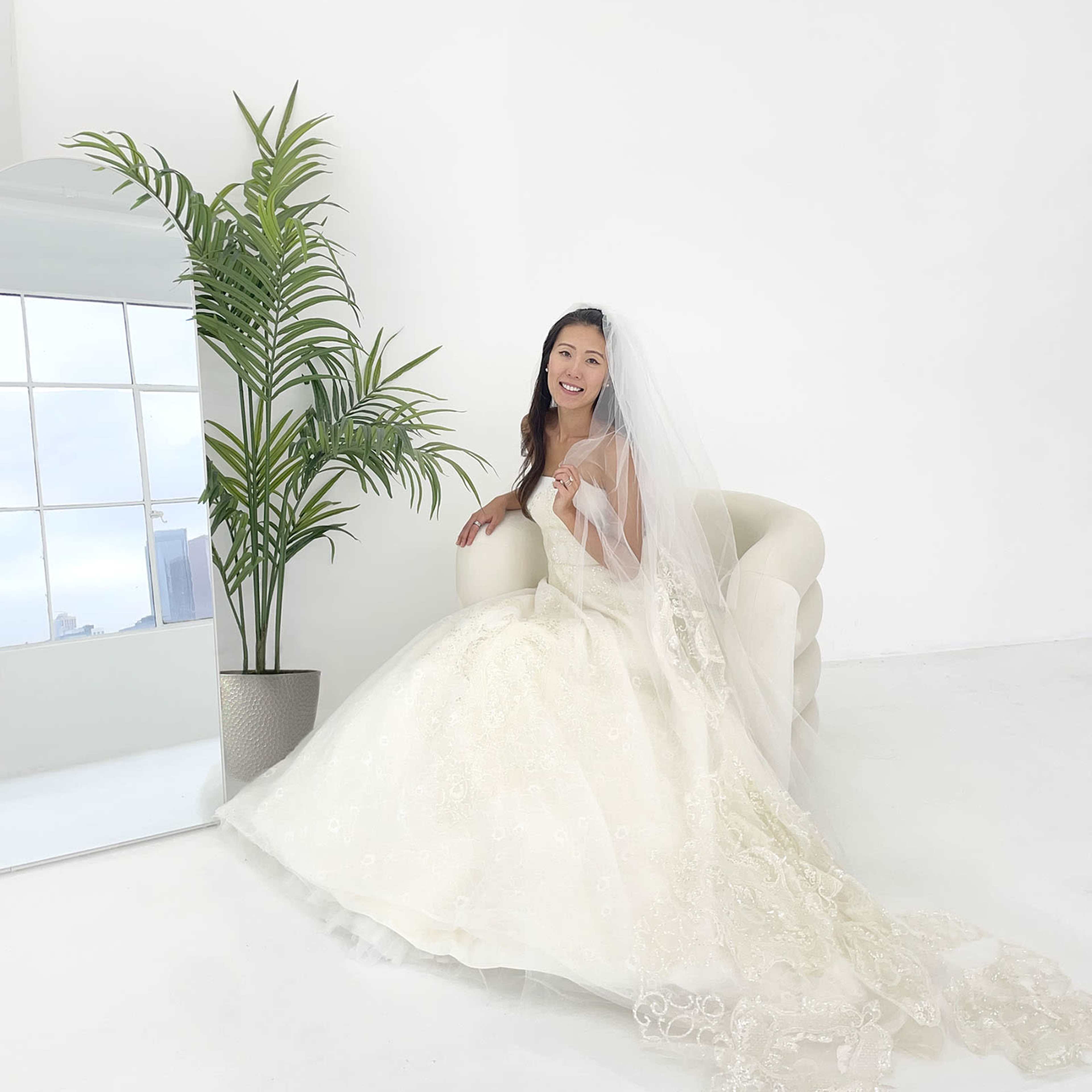 A woman in a white wedding dress and veil sits on a chair beside a tall plant and a large mirror against a plain background.