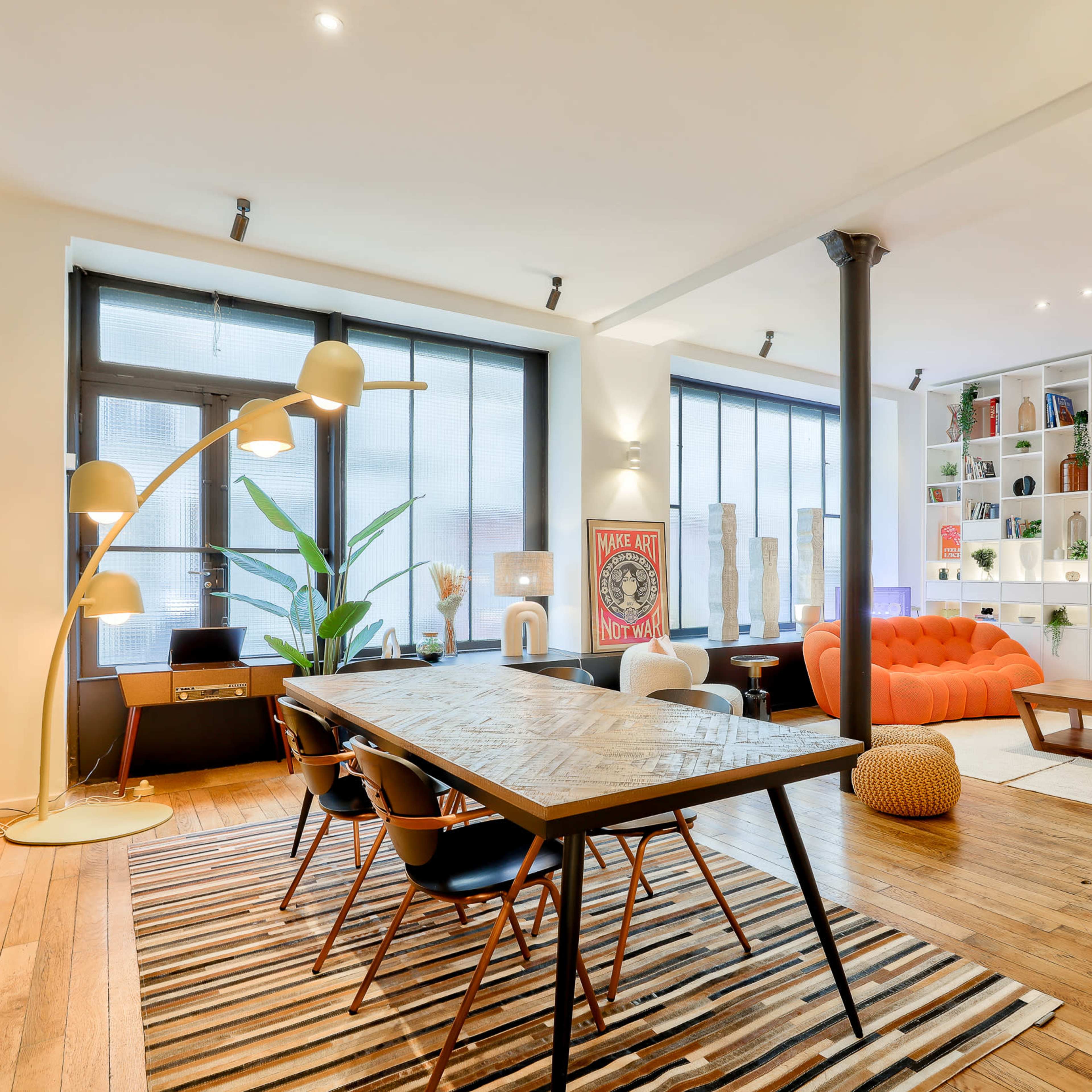 A modern living space features a large orange sofa, a dining table with black chairs, and shelves filled with decorative items, illuminated by natural light from large windows.