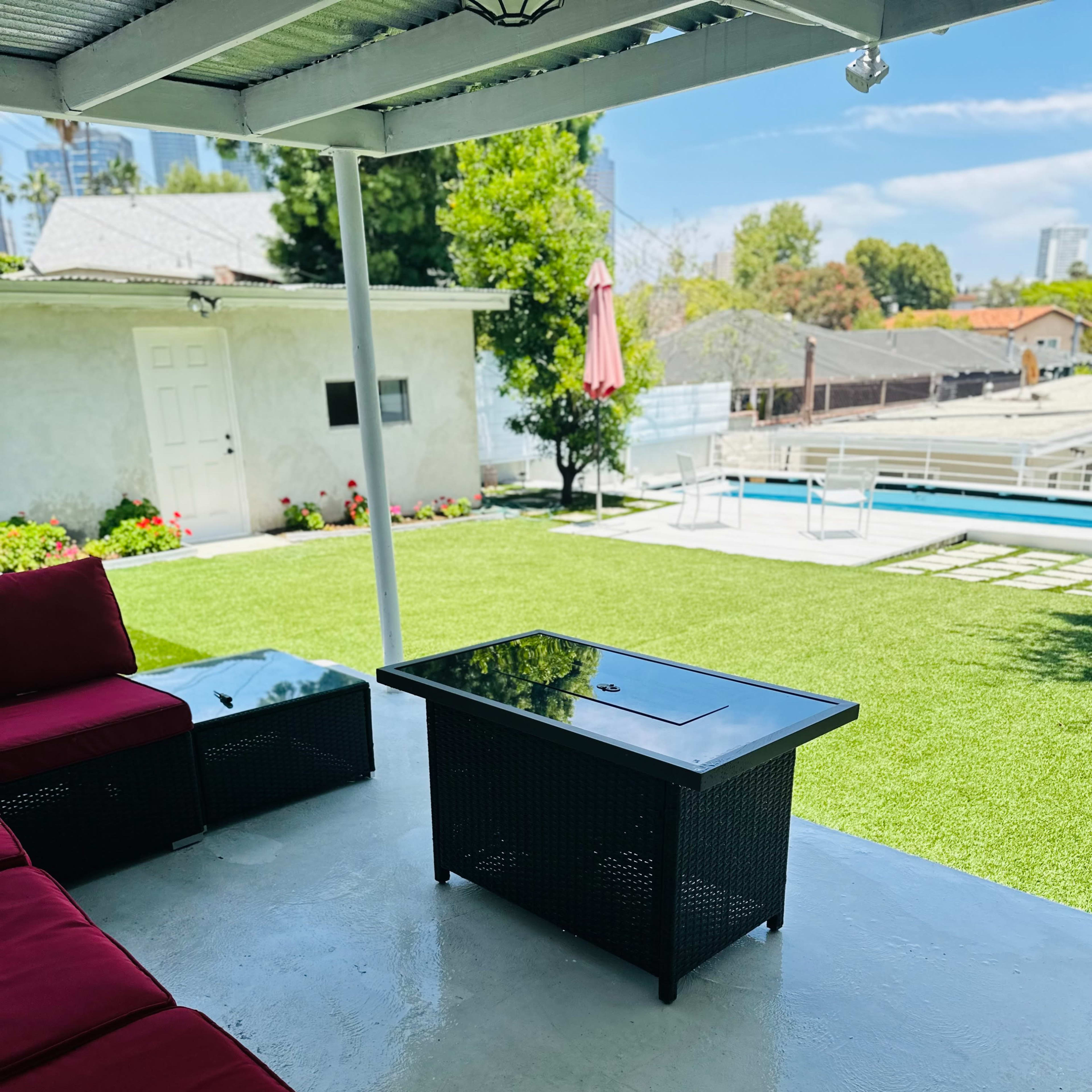 A covered outdoor seating area features red couches and a glass-top coffee table overlooking a green lawn and a swimming pool.