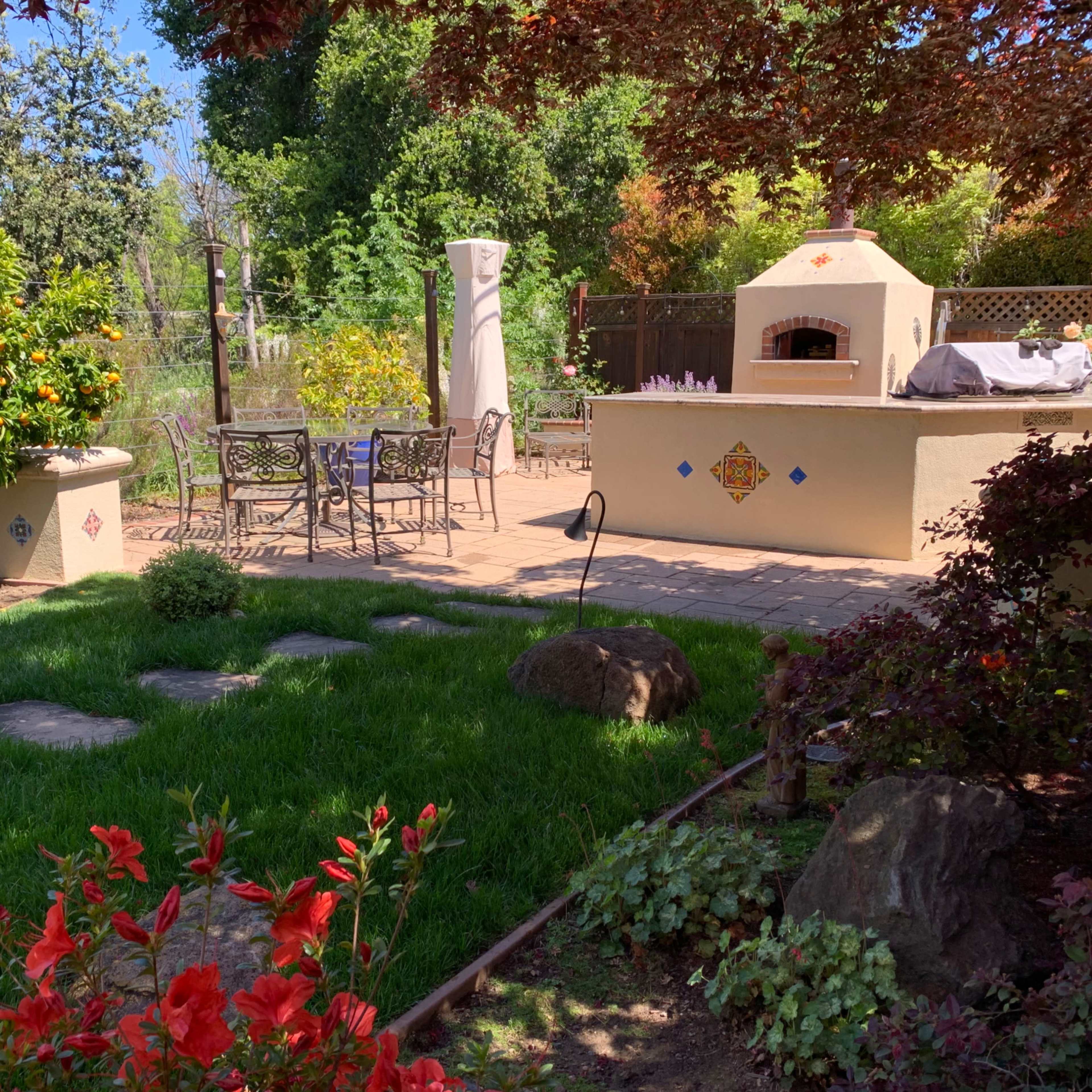A landscaped backyard with a stone patio, an outdoor kitchen, a dining set, and various plants and trees.