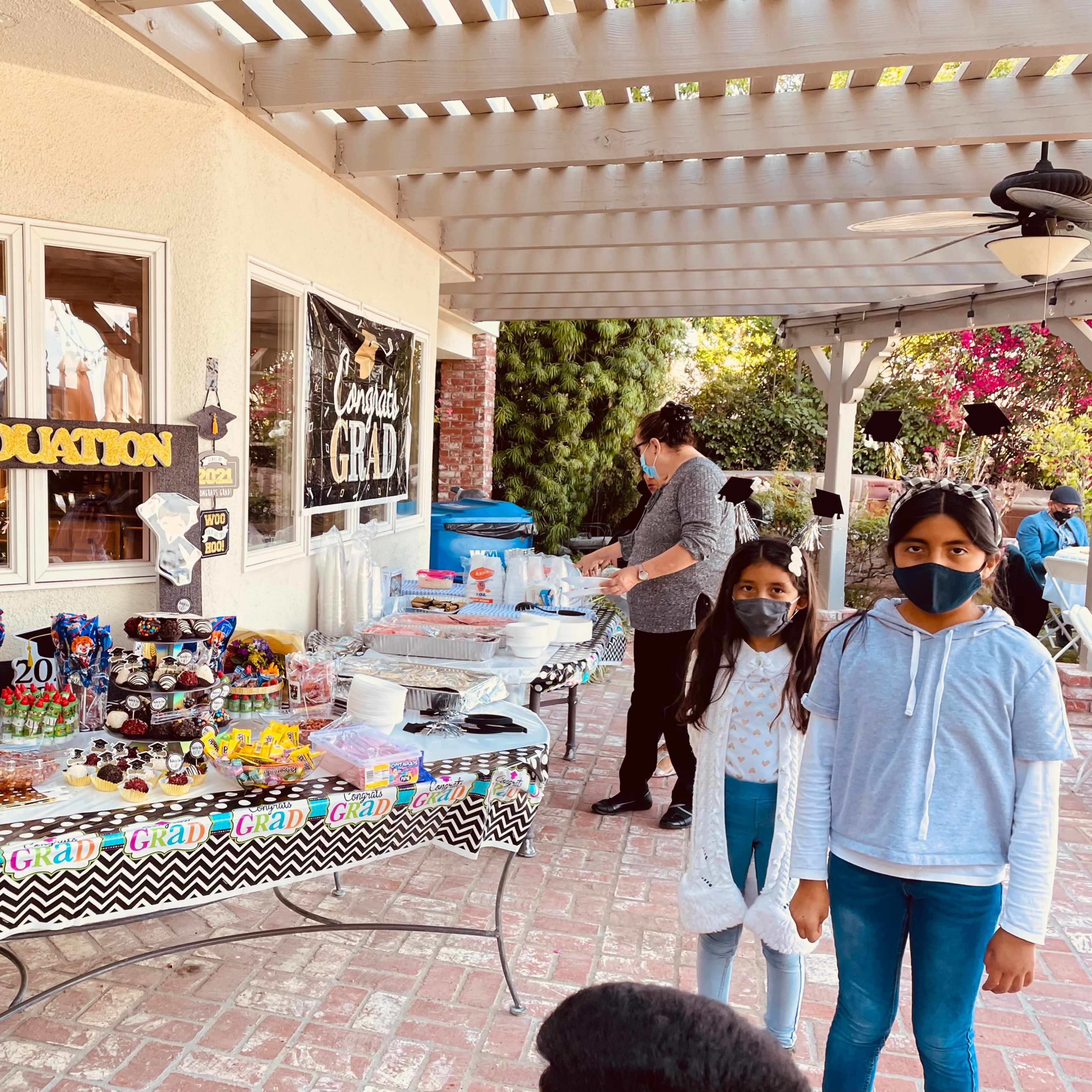 A graduation celebration is set up outdoors with a decorated table displaying snacks and desserts, while two girls in masks pose in front.