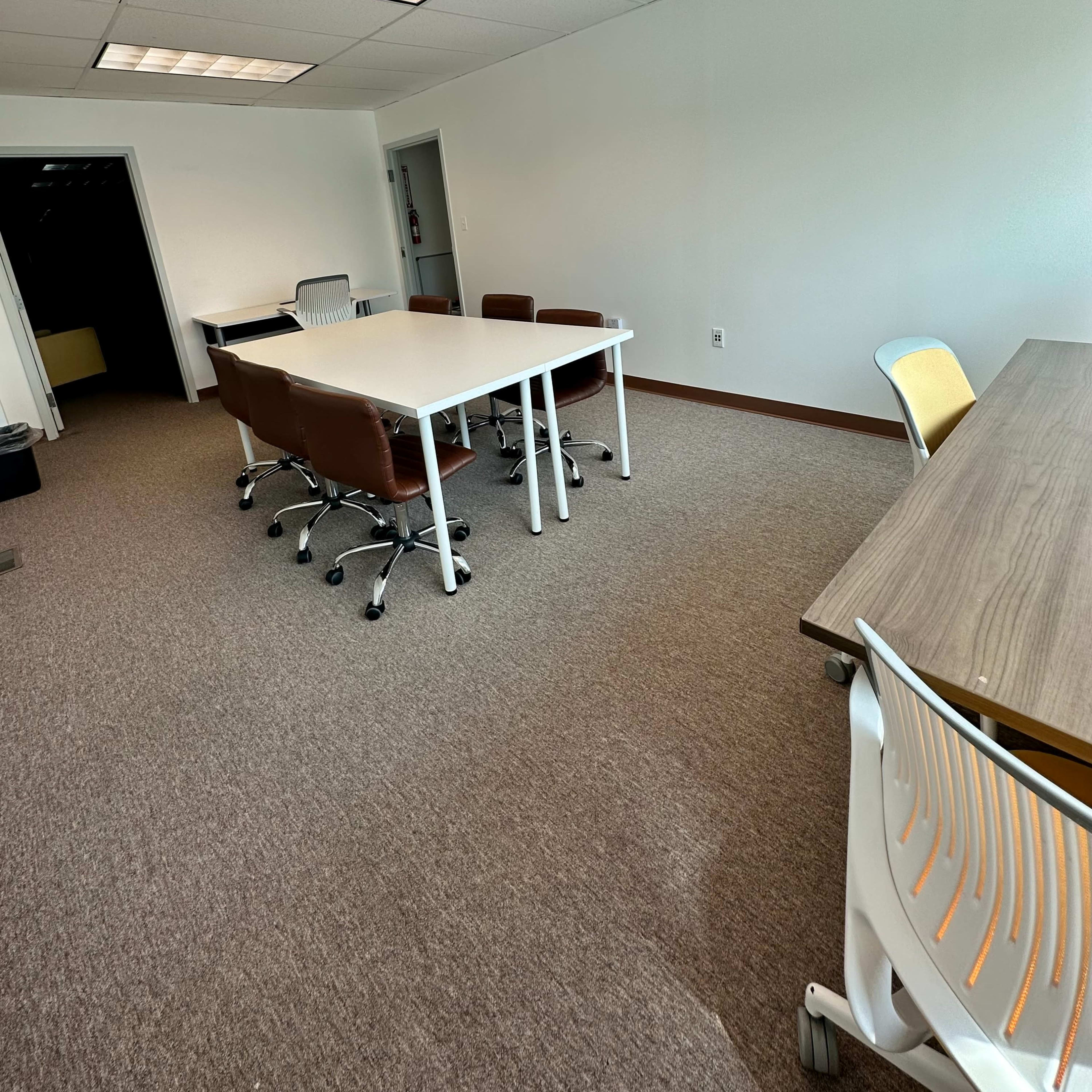 A spacious office meeting room with a white table surrounded by brown chairs and a wooden table against the wall.