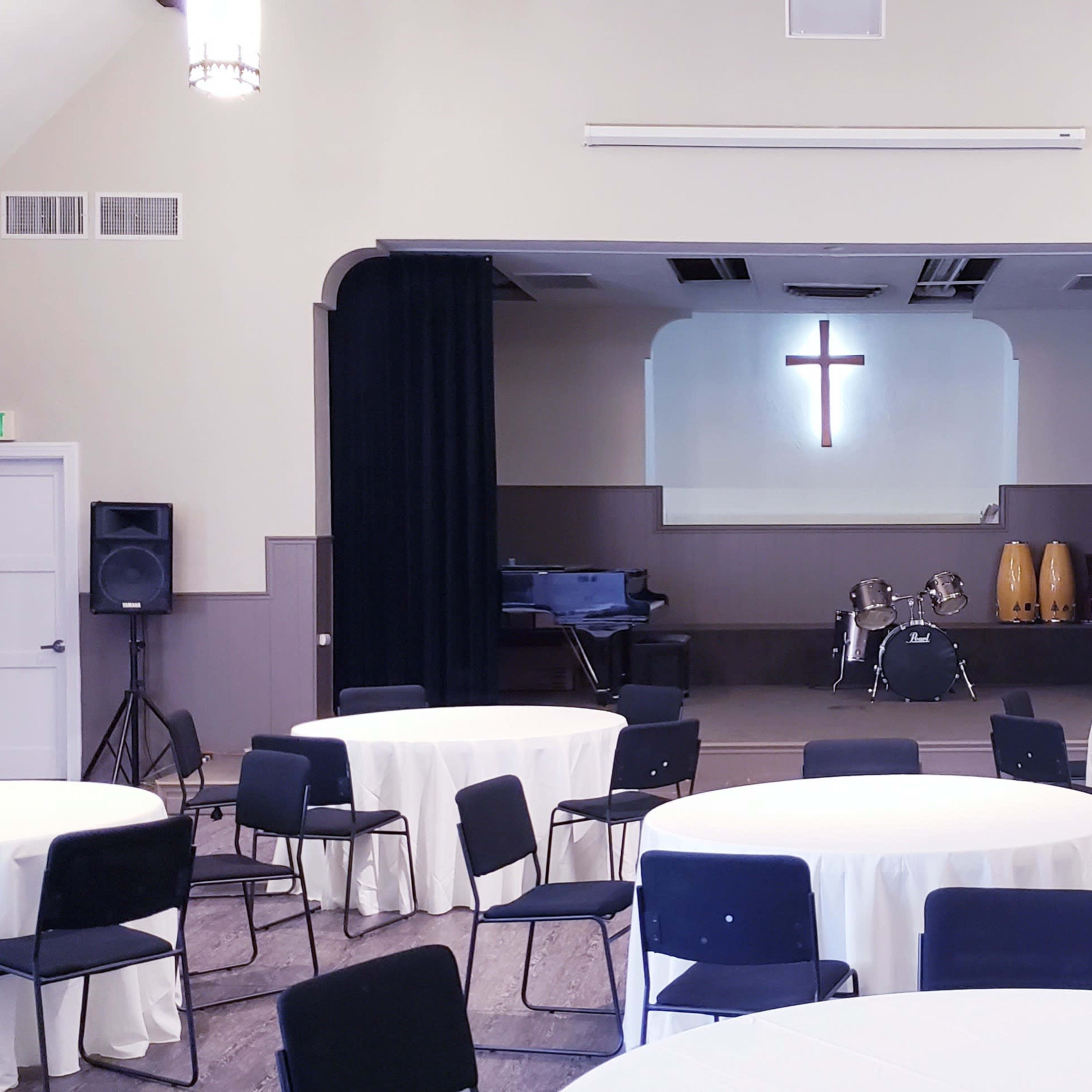 A church hall set up with round tables and chairs arranged before a stage featuring a cross and musical instruments.