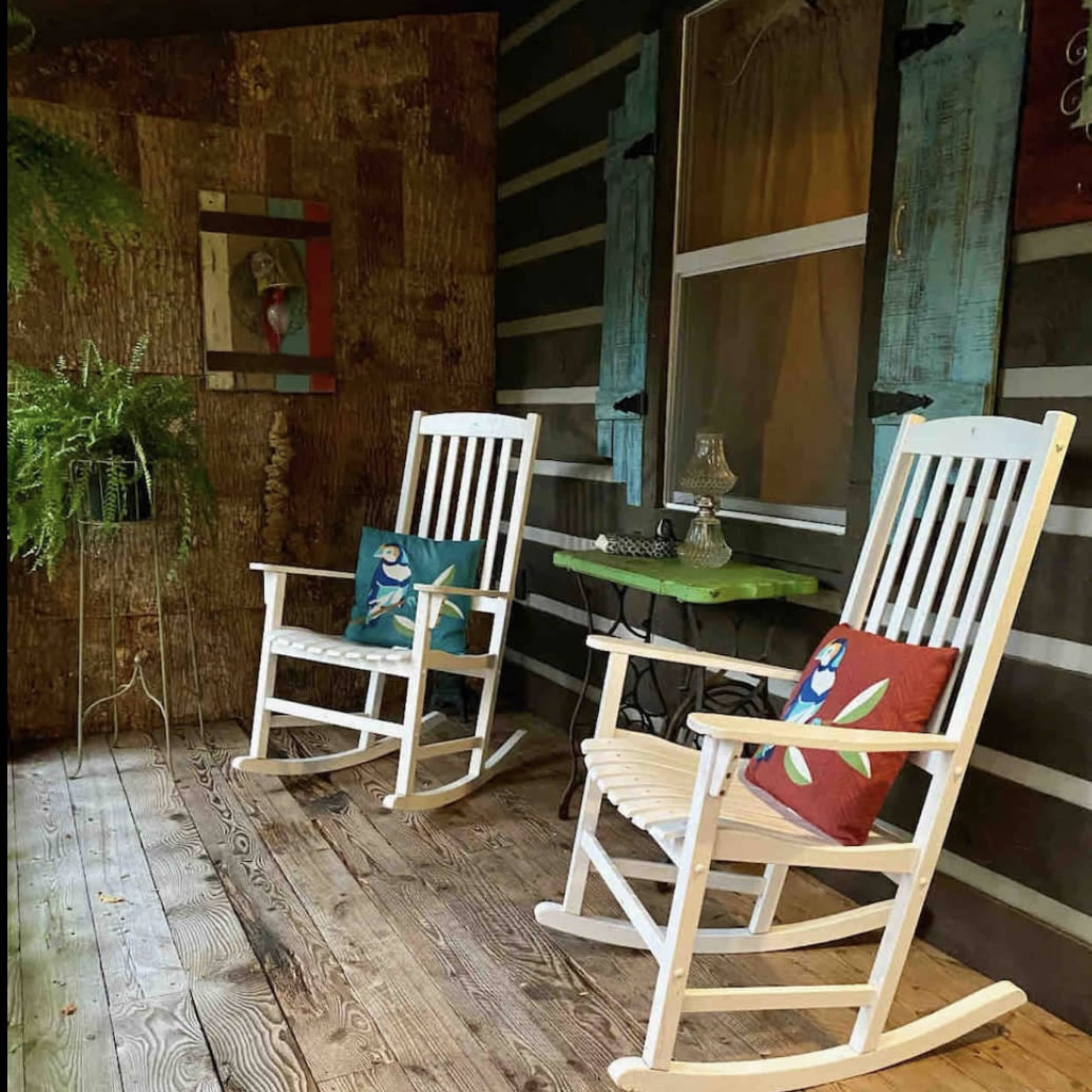 Two white rocking chairs with decorative cushions placed on a wooden porch beside a small green table and a fern.