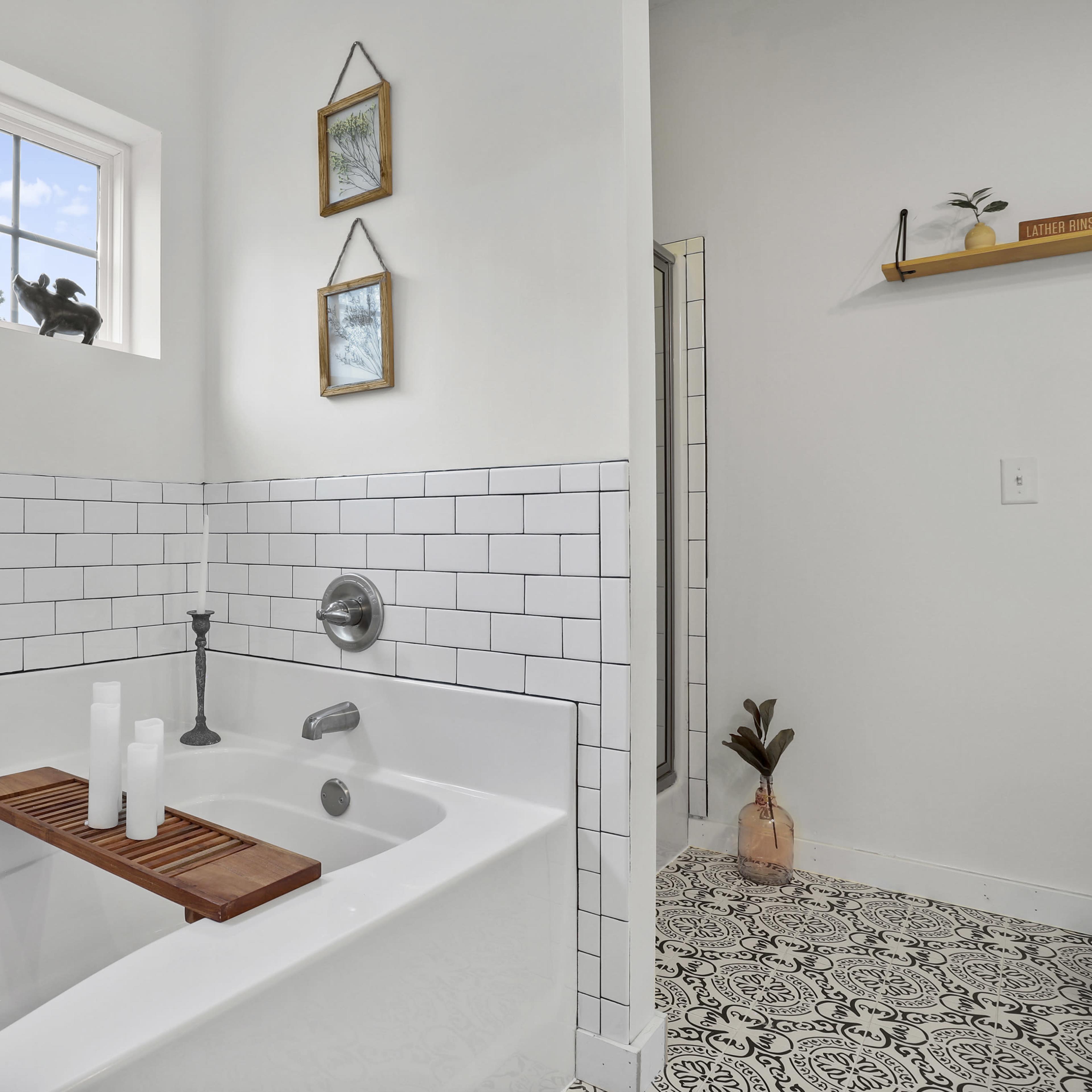 A bathroom featuring a white bathtub with a wooden tray, white tiled walls, and a patterned floor, with a small shower area visible in the background.