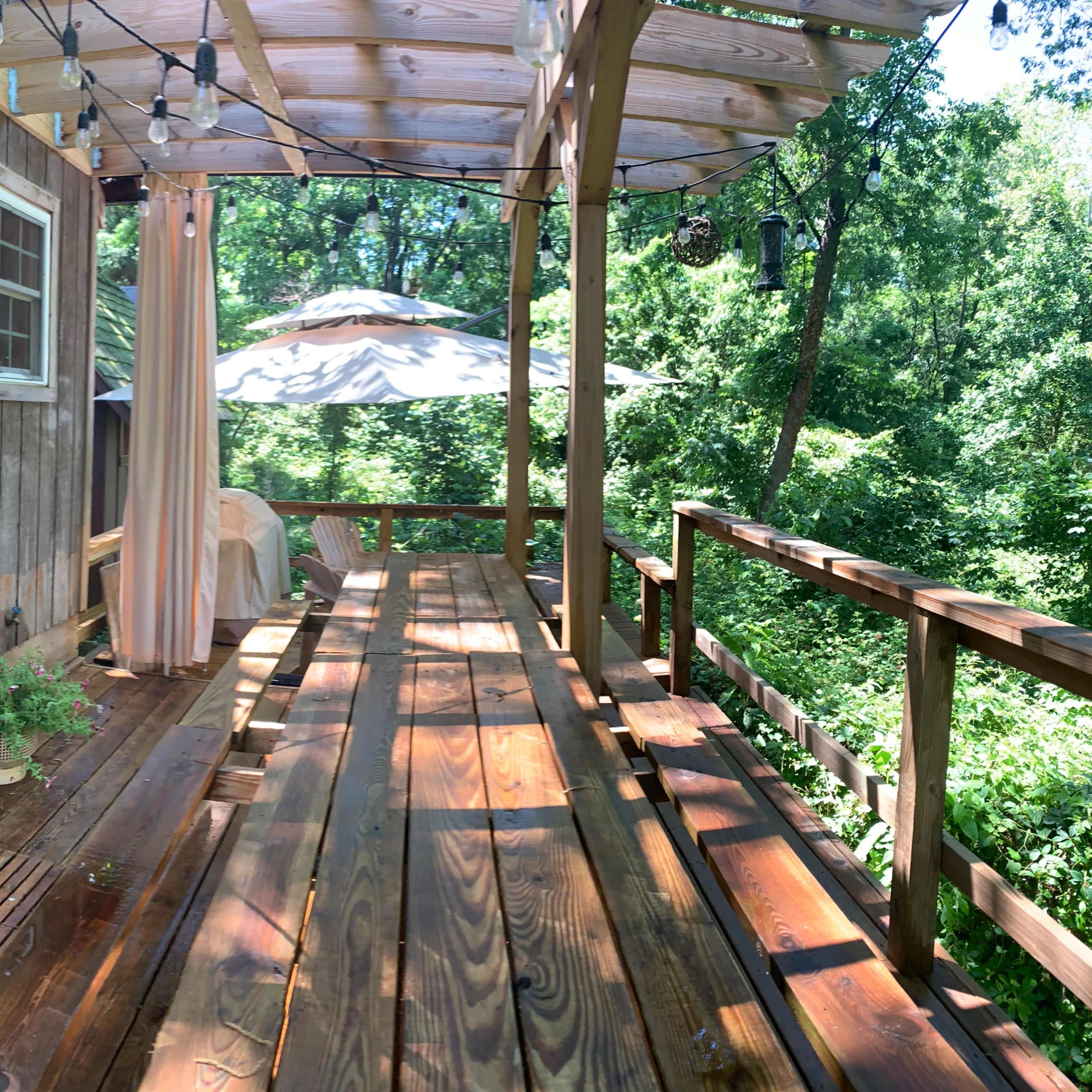 A wooden deck with a parasol, surrounded by trees and featuring string lights and a potted plant.