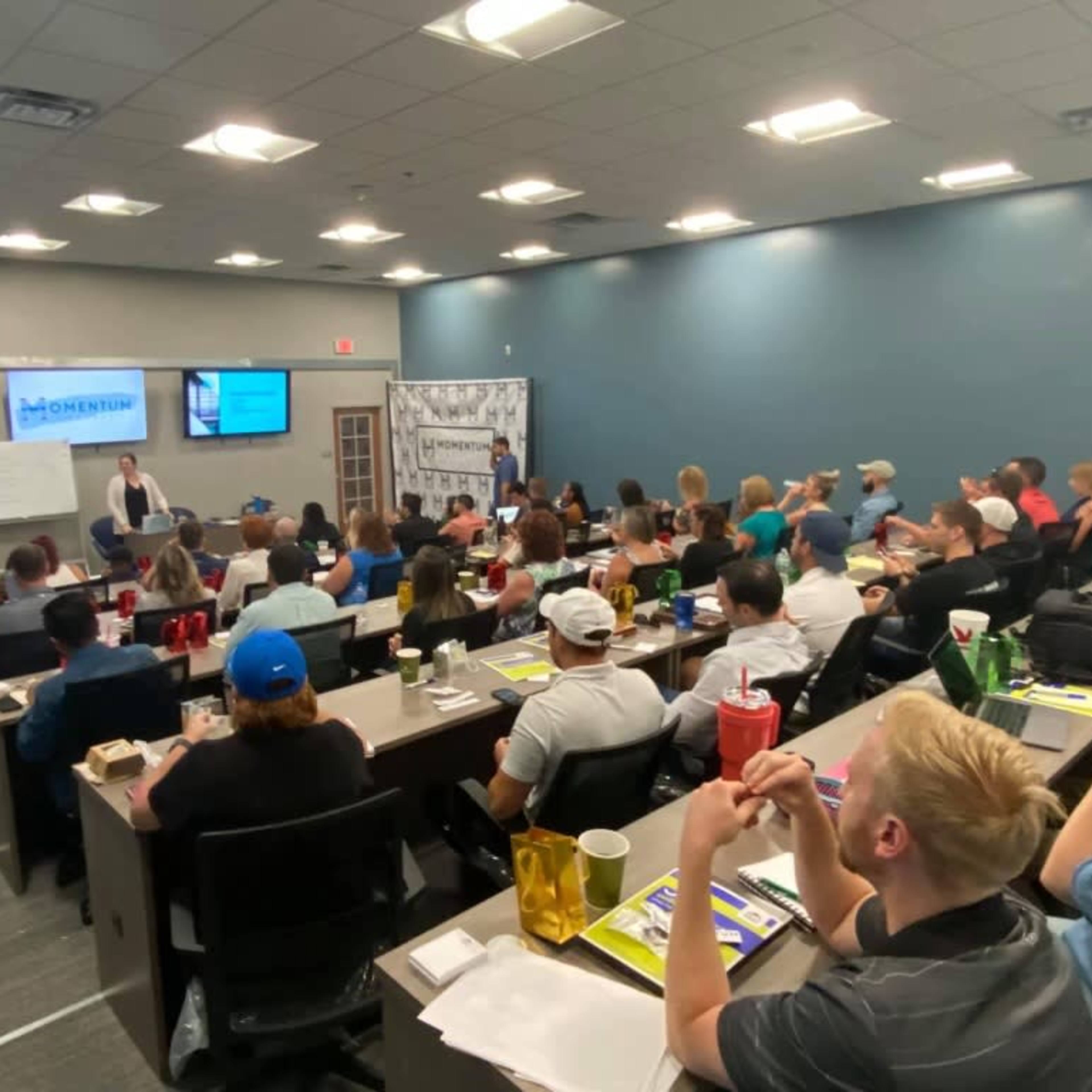 A classroom filled with attendees participates in a presentation with a speaker at the front and a projector displaying information.