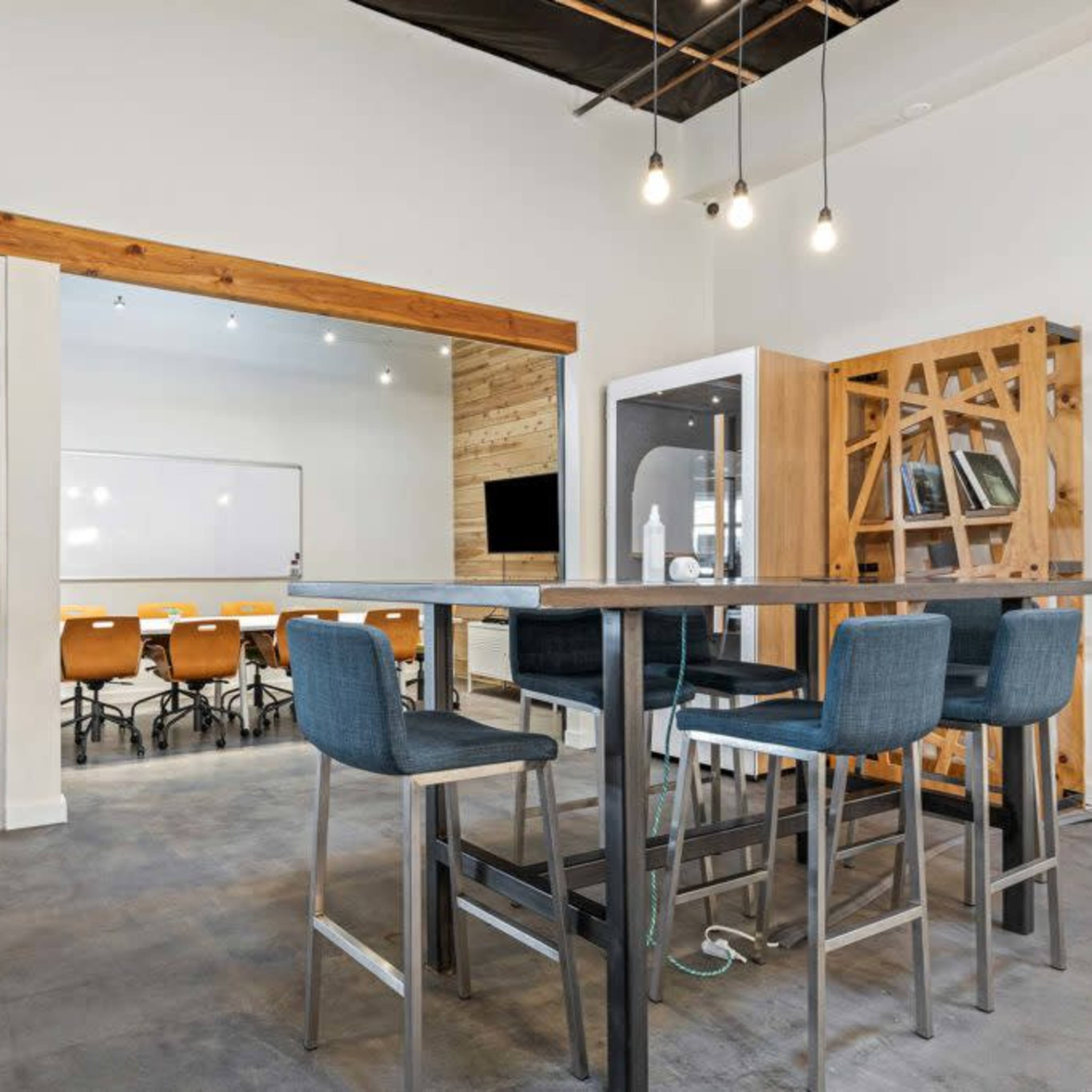 A modern office space features a high table with four chairs, a glass meeting room, and a large whiteboard in the background.