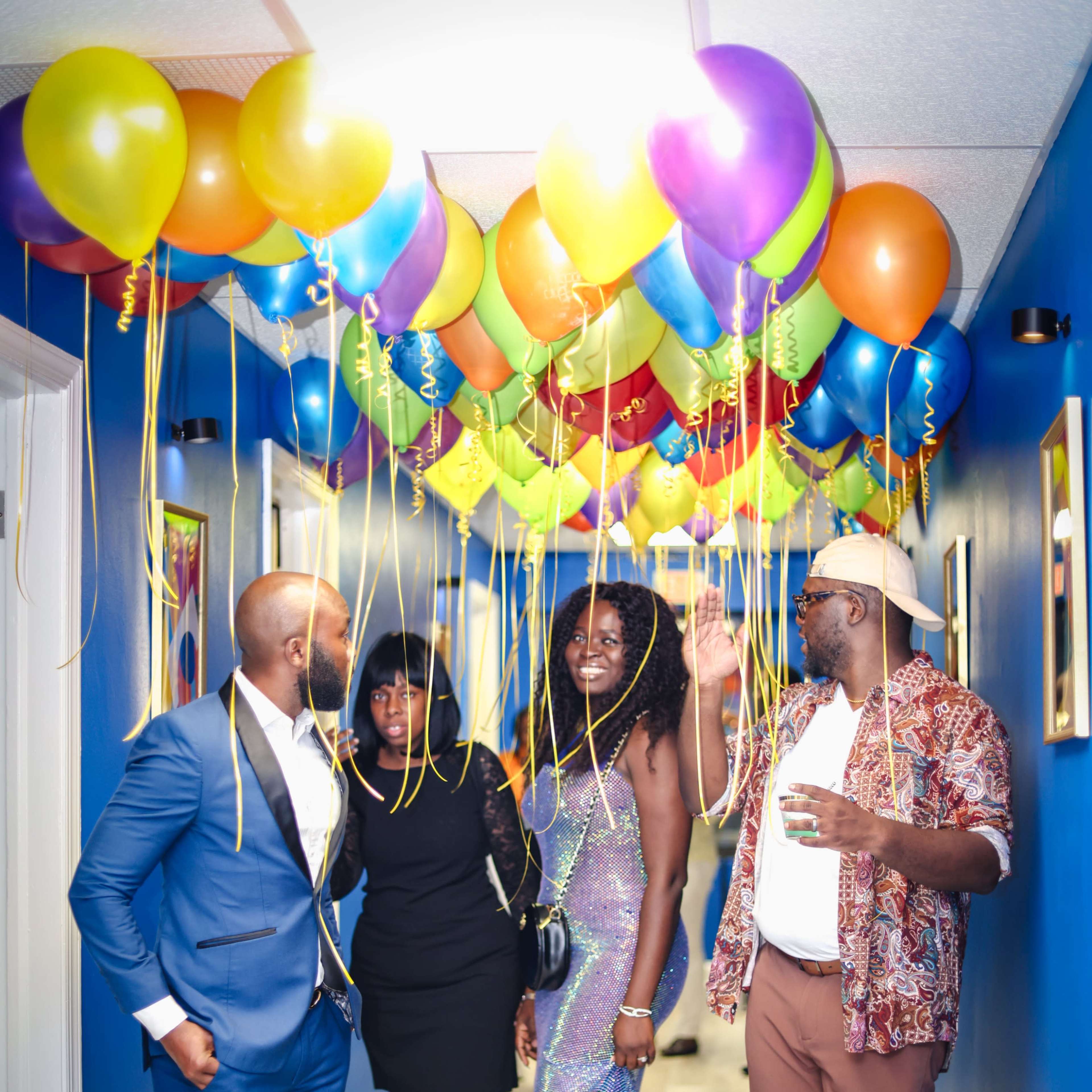 A group of four people stands in a hallway decorated with colorful balloons hanging from above.