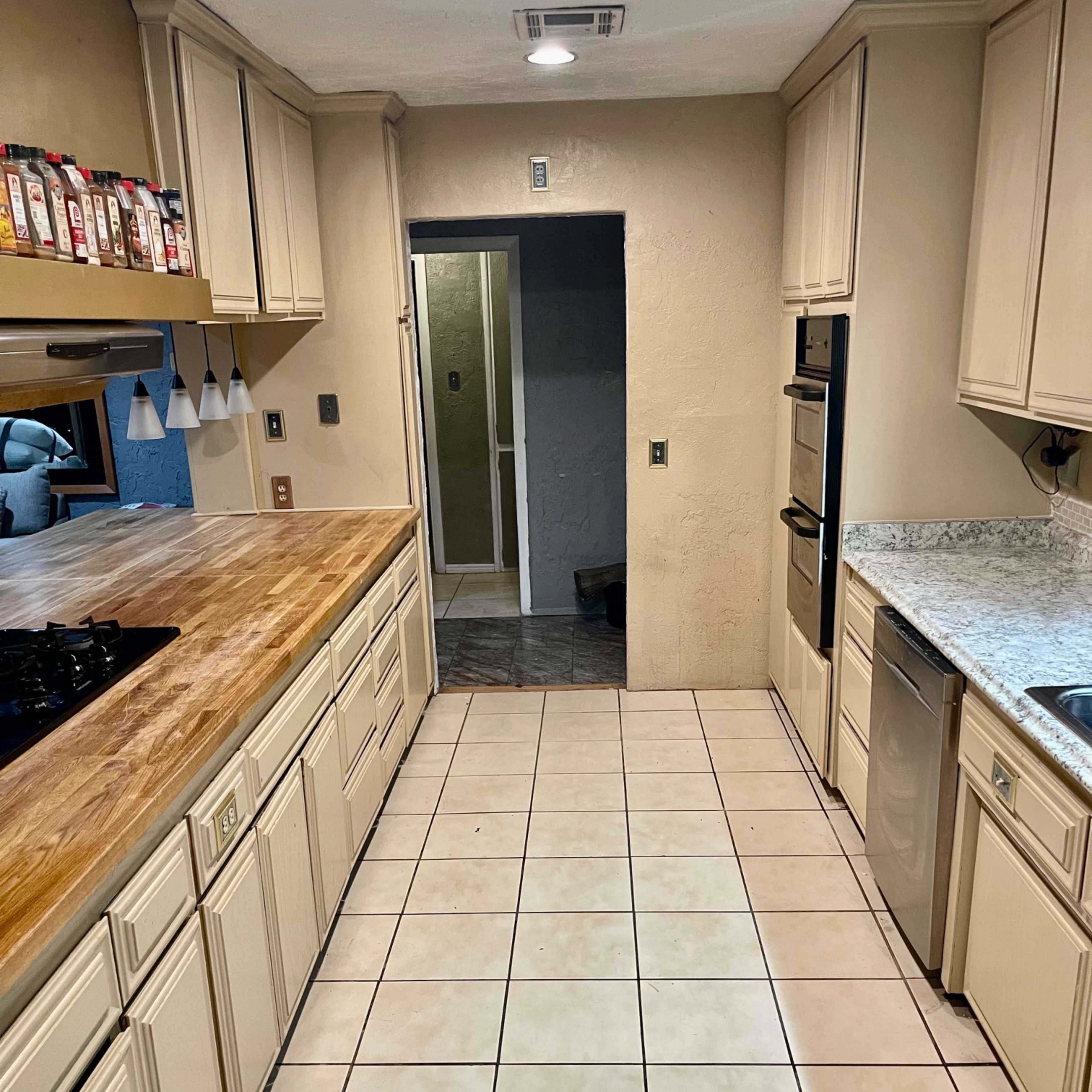 A kitchen with beige cabinets, a wooden countertop, stainless steel appliances, and tiled flooring, leading to an entryway with a bathroom visible in the background.