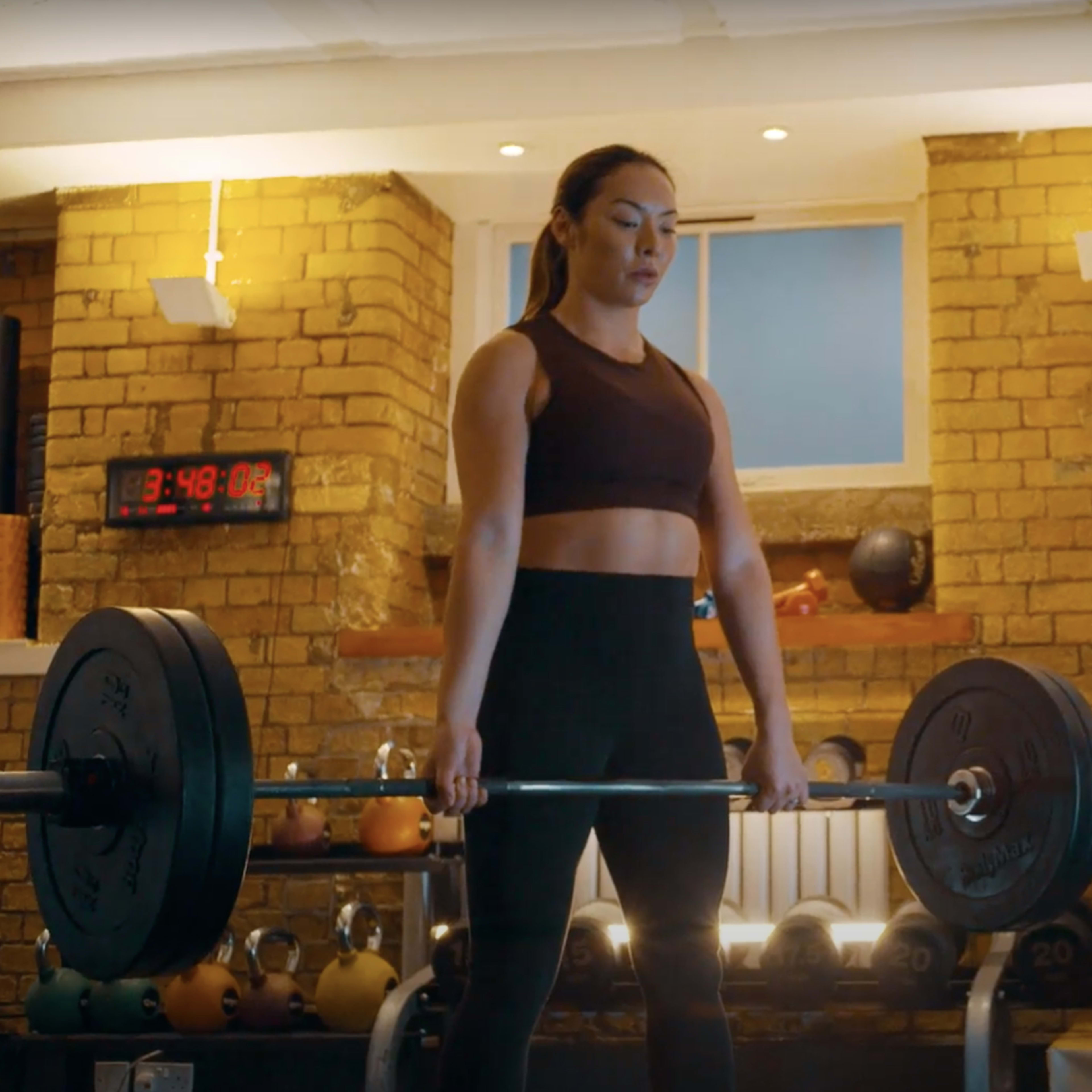 Two women are in a gym setting, with one lifting a barbell while the other observes from the side.