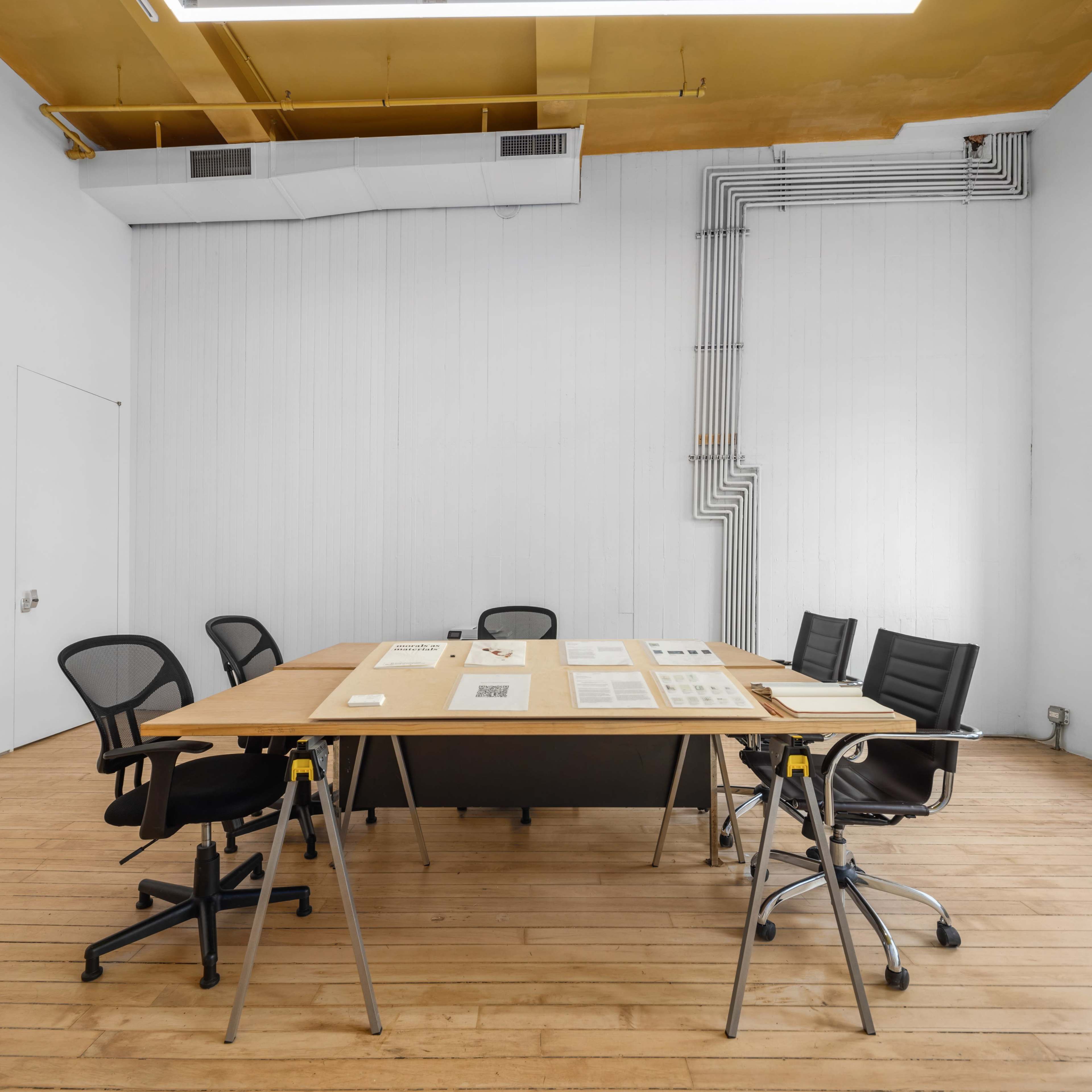 A conference room features a large wooden table surrounded by black office chairs and a plain white wall with visible electrical conduits.