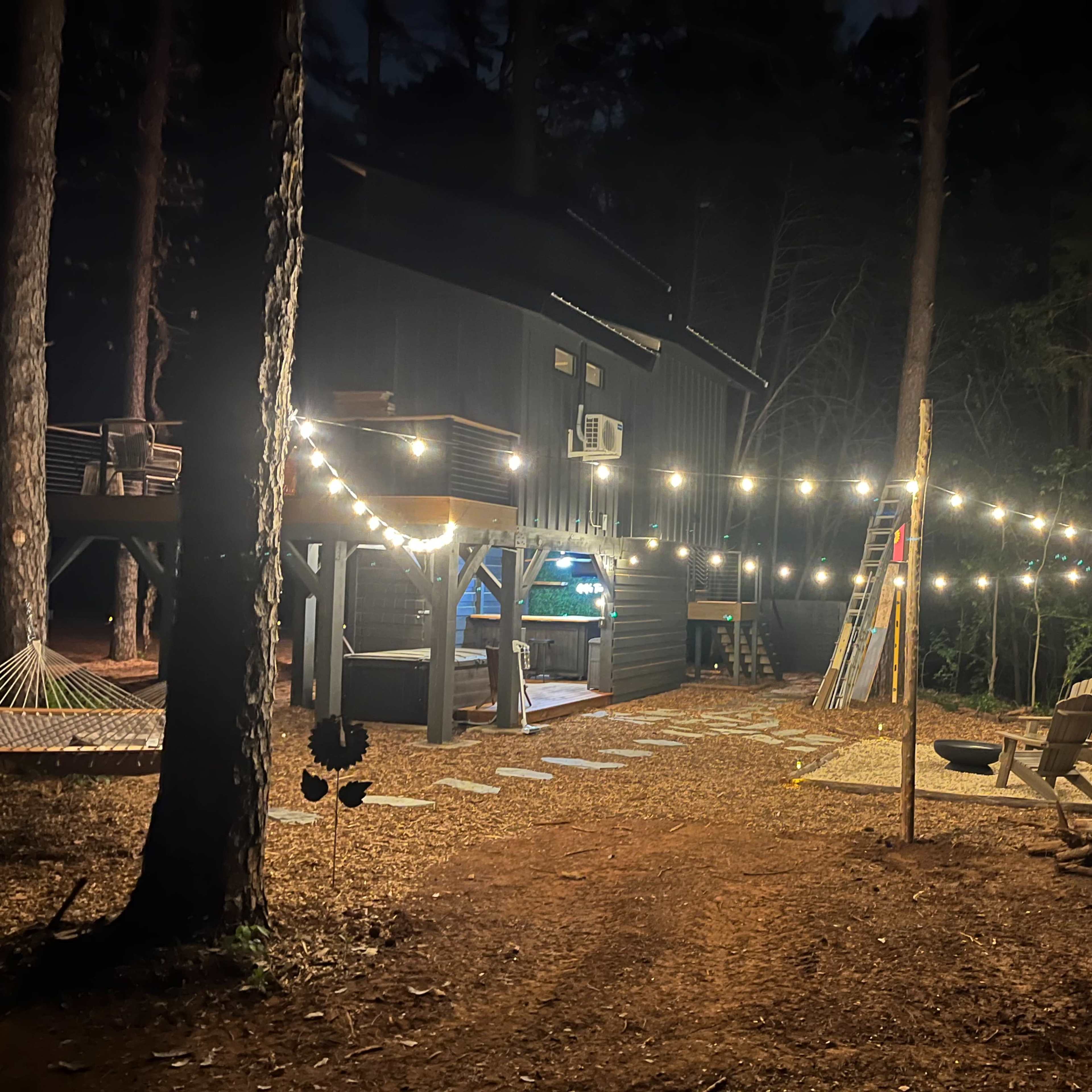 A dark outdoor area illuminated by string lights, with a wooden building, a hammock, and a fire pit surrounded by pine trees.