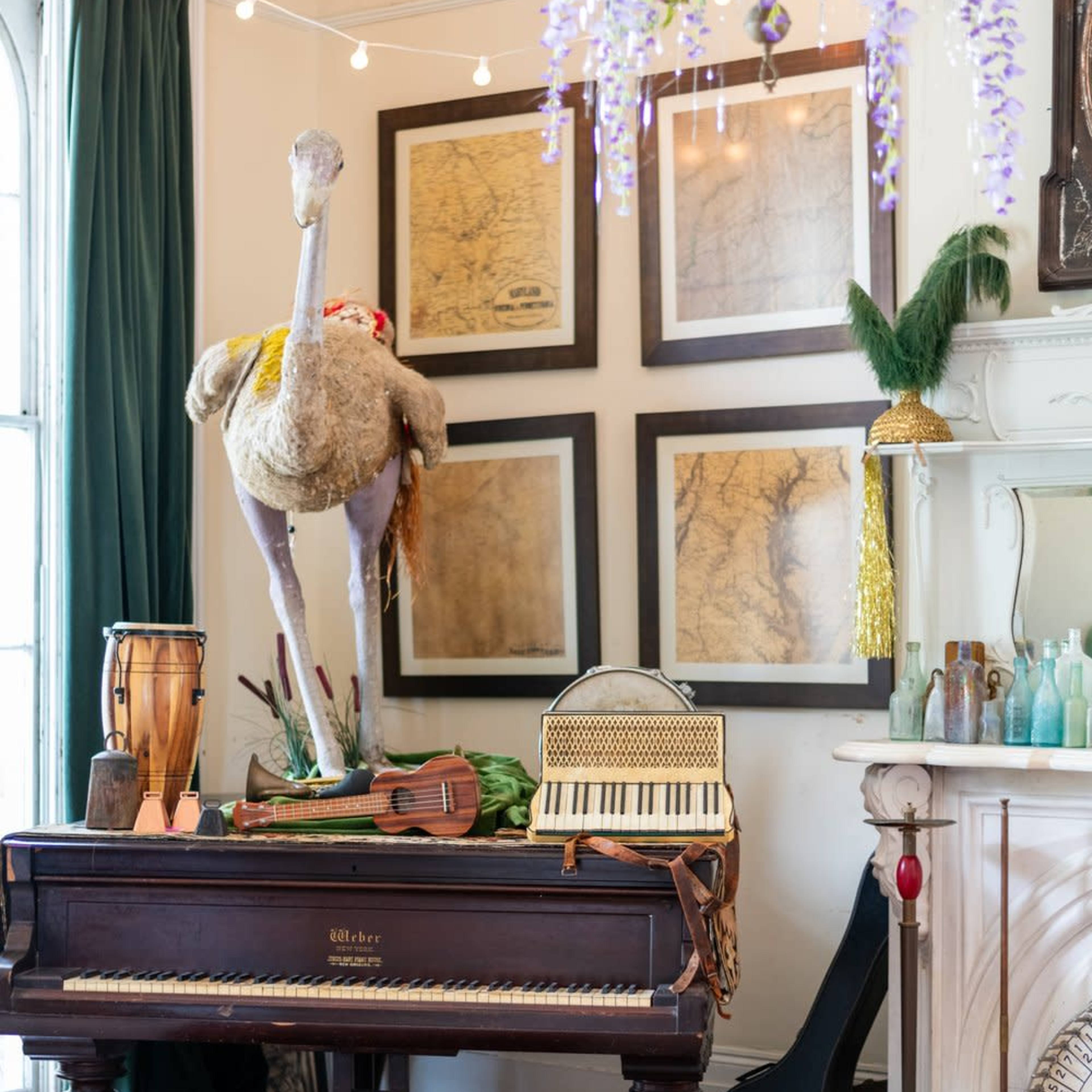 A vintage piano with a red bench is positioned near an ornate fireplace, accompanied by an oversized stuffed ostrich and various musical instruments, surrounded by framed maps and decorative elements.