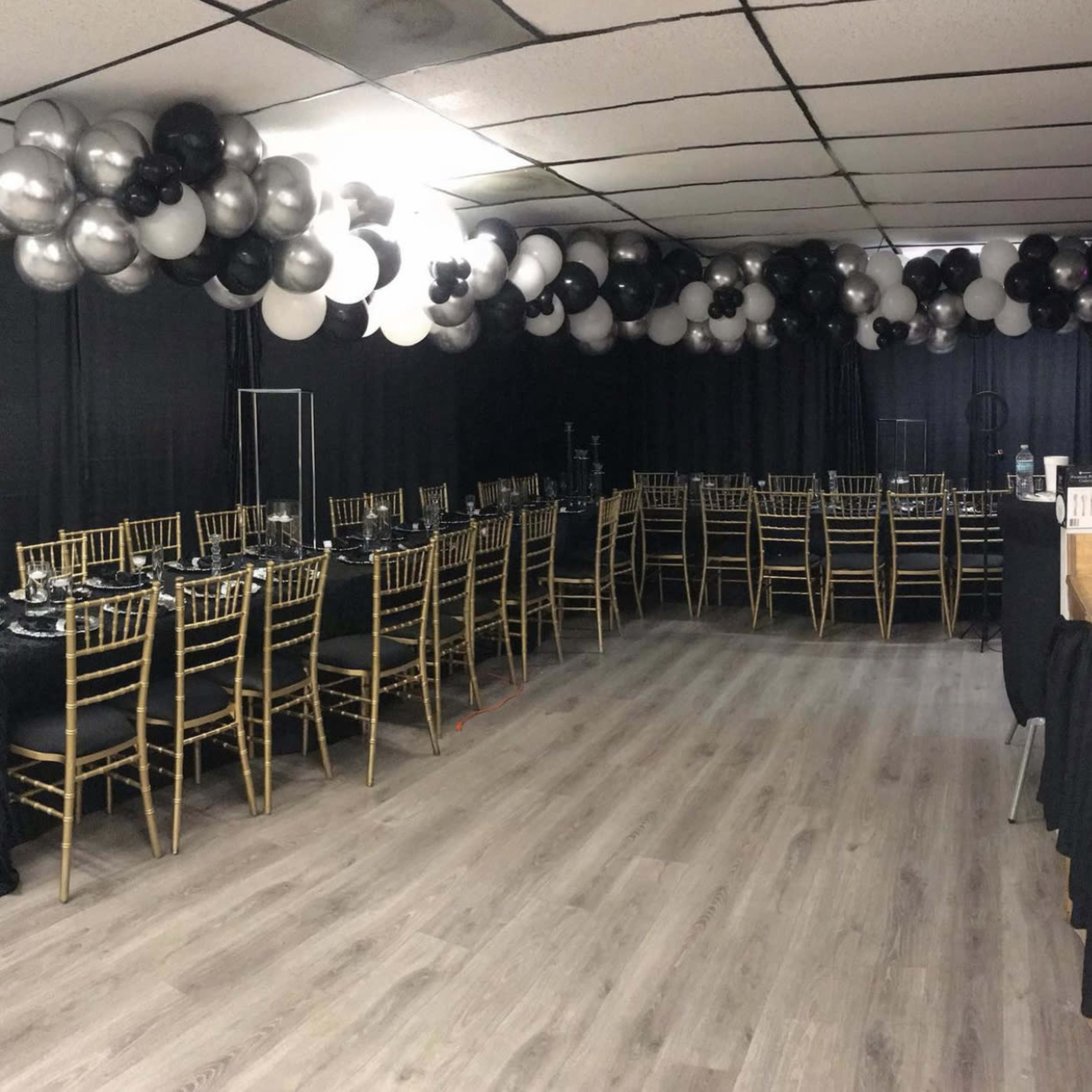 A dining area is set up with multiple rows of gold Chiavari chairs and black tables, decorated with silver and black balloons overhead.