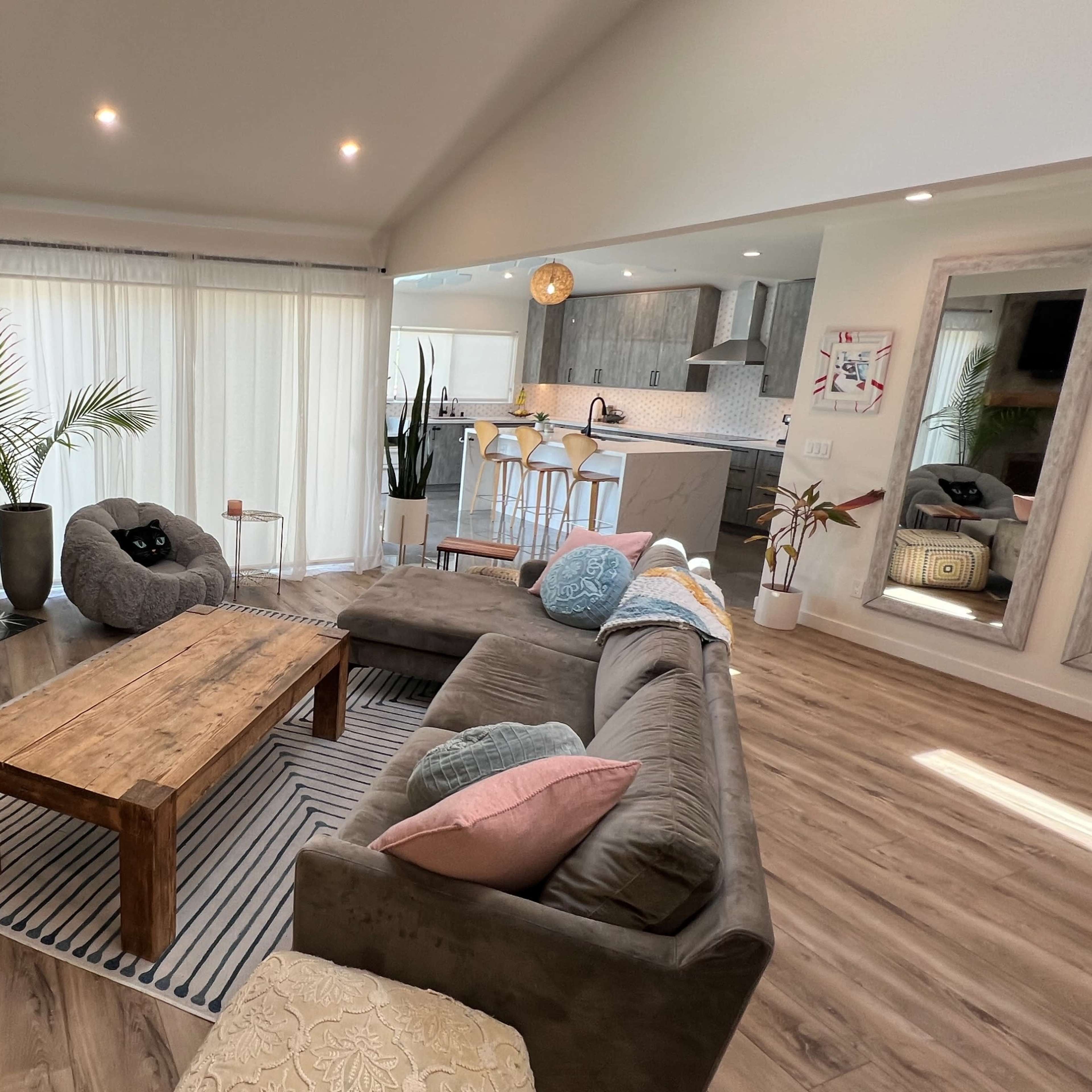 A modern living room with a sectional sofa, a wooden coffee table, a television mounted on the wall, and large windows displaying natural light.
