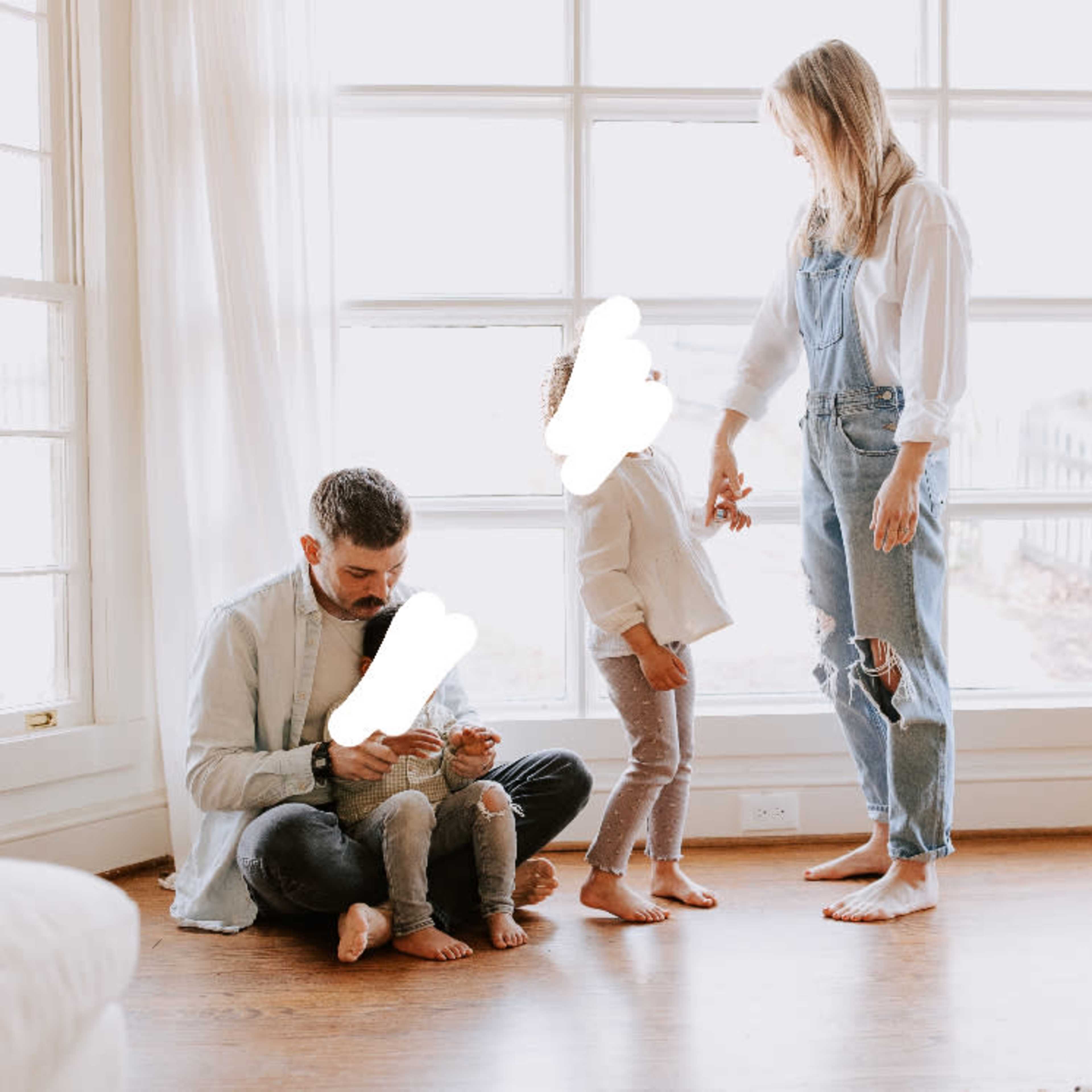 A family sits in a sunlit living room, with one parent on the floor using a phone, a child standing beside them, and the other parent interacting while a dog watches.