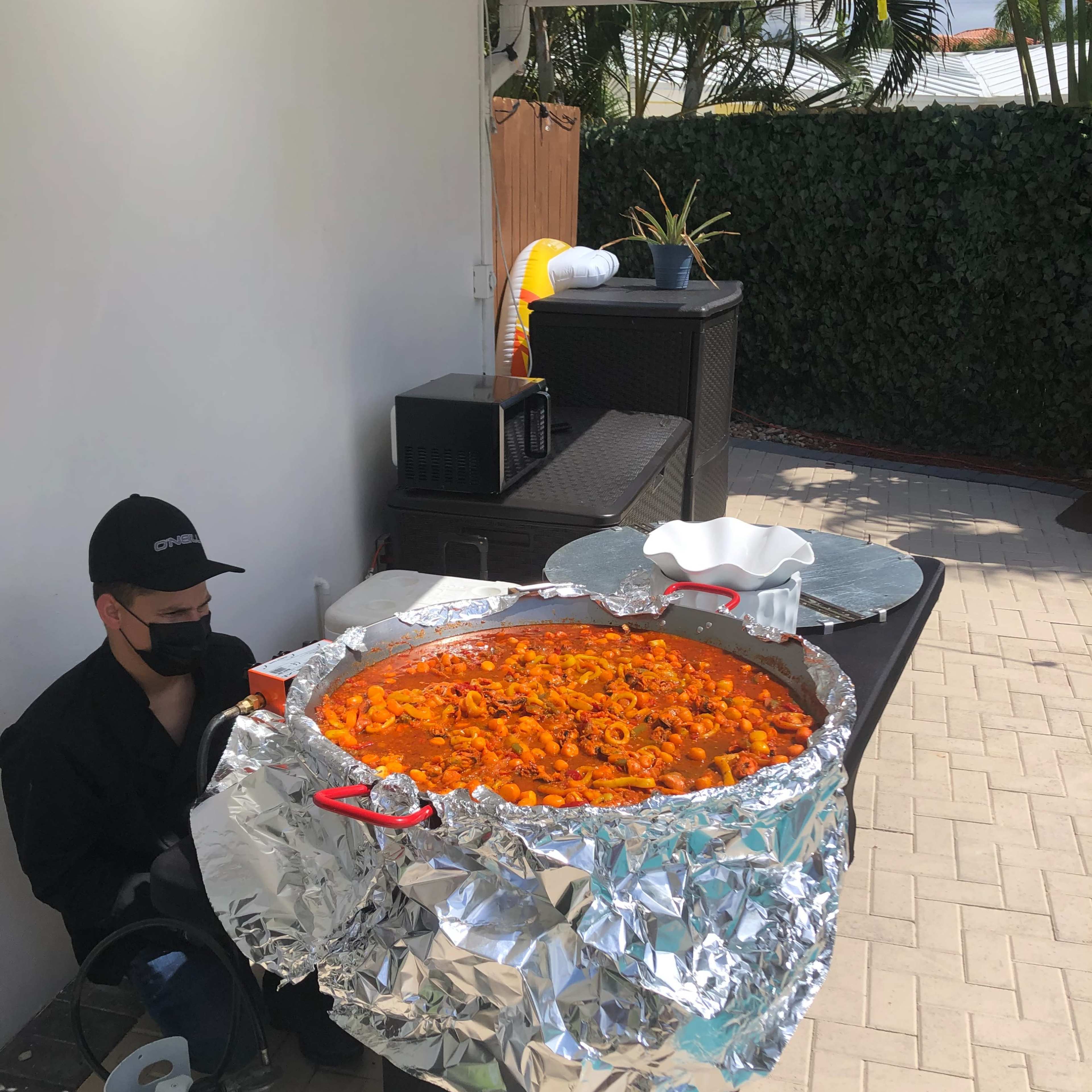 A person in a black hat and mask stands beside a large pot of food covered in aluminum foil on a table outdoors.
