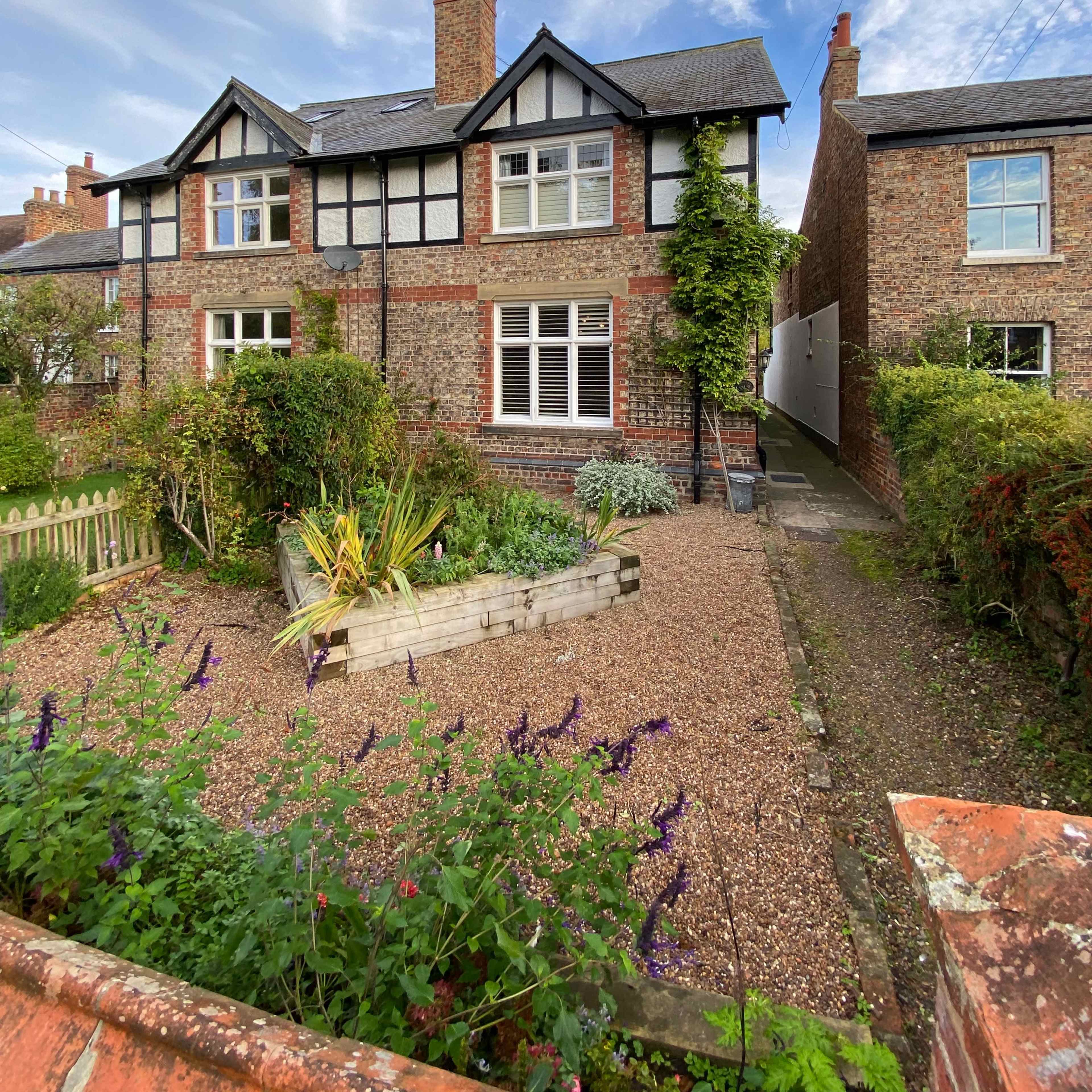 A two-story brick house with a tiled roof is surrounded by a gravel garden and a fence, featuring a variety of plants and shrubs.