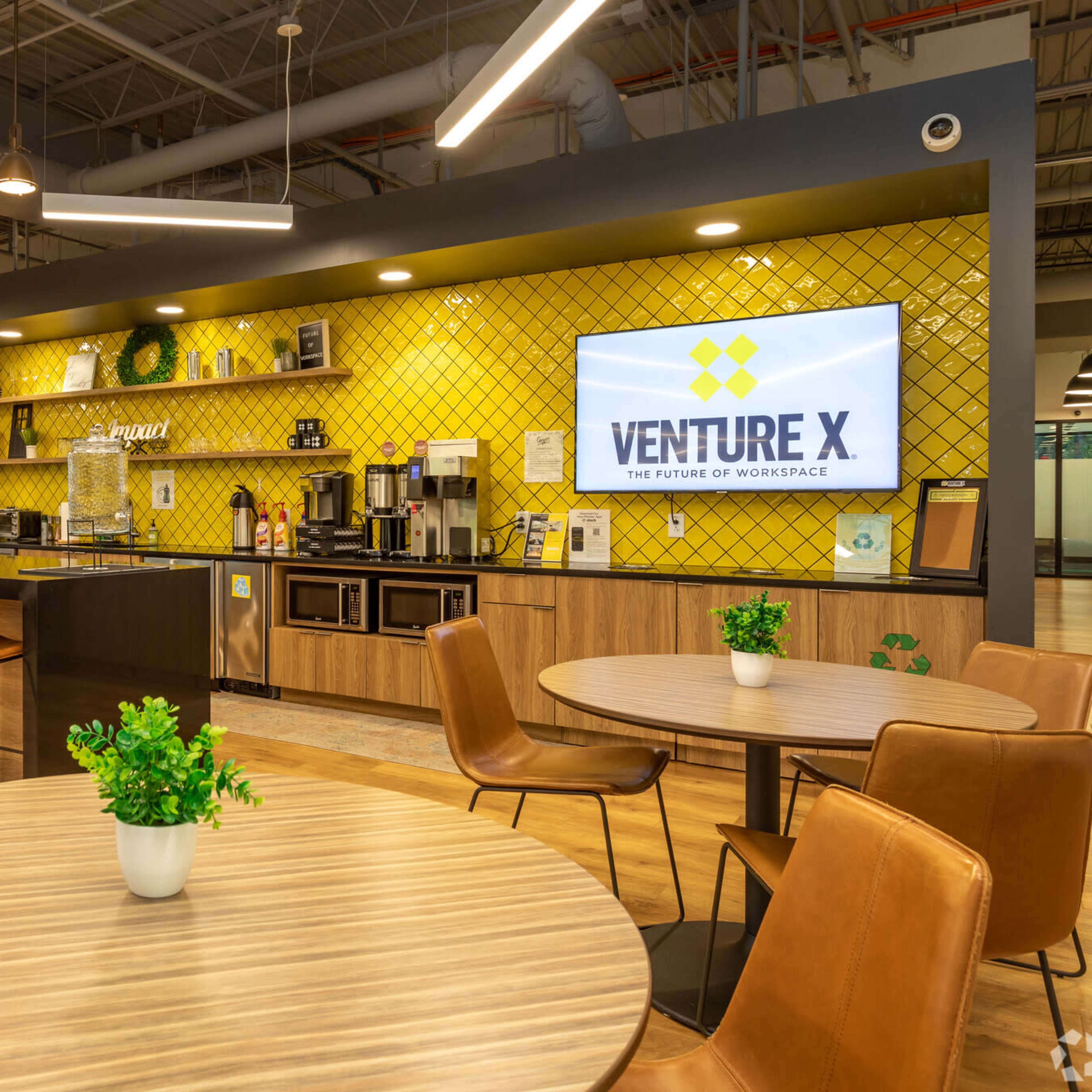 A modern coworking space featuring a bar area with yellow tiled walls, seating at a counter, and tables with plants.