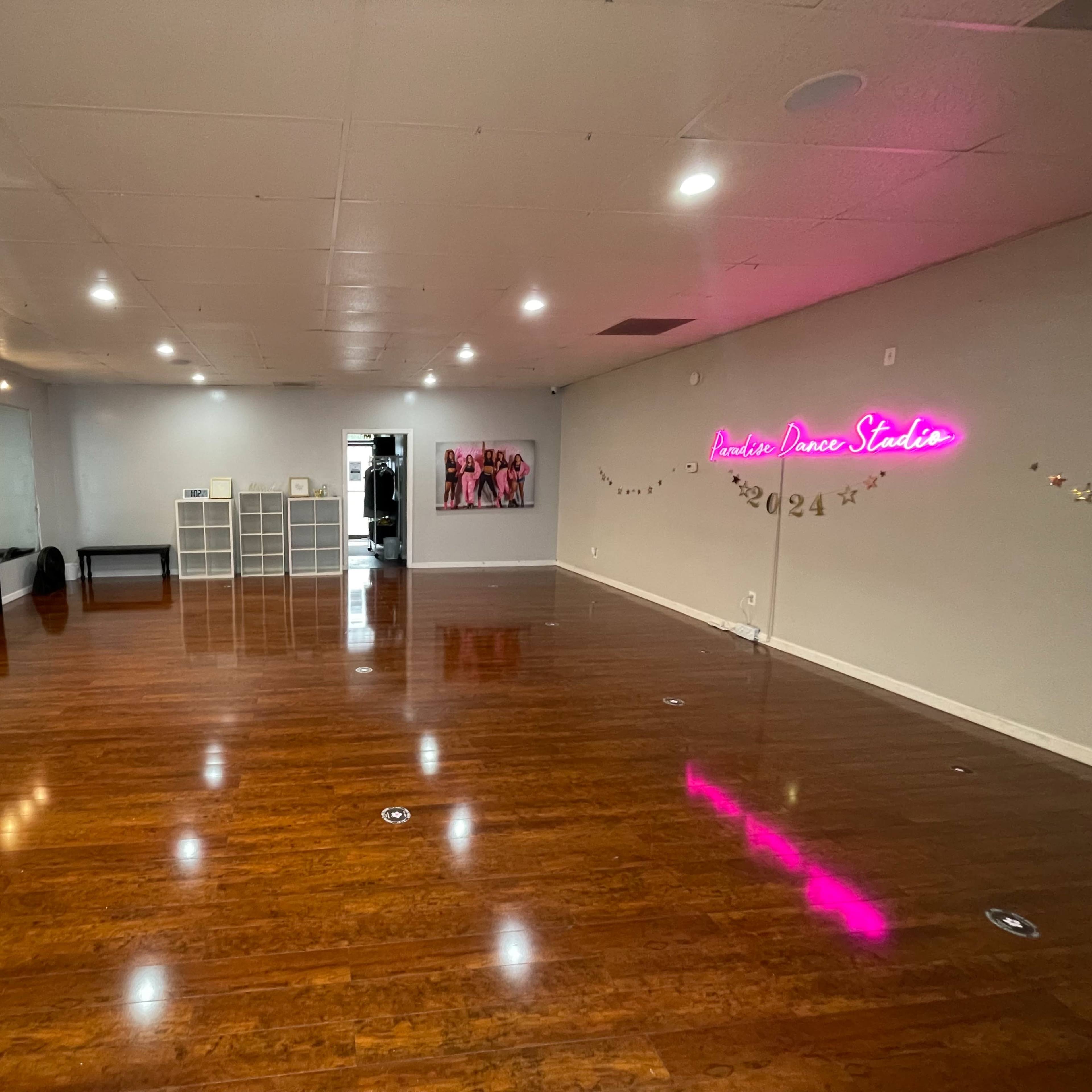 An empty dance studio with wooden flooring, wall mirrors, and a neon sign.