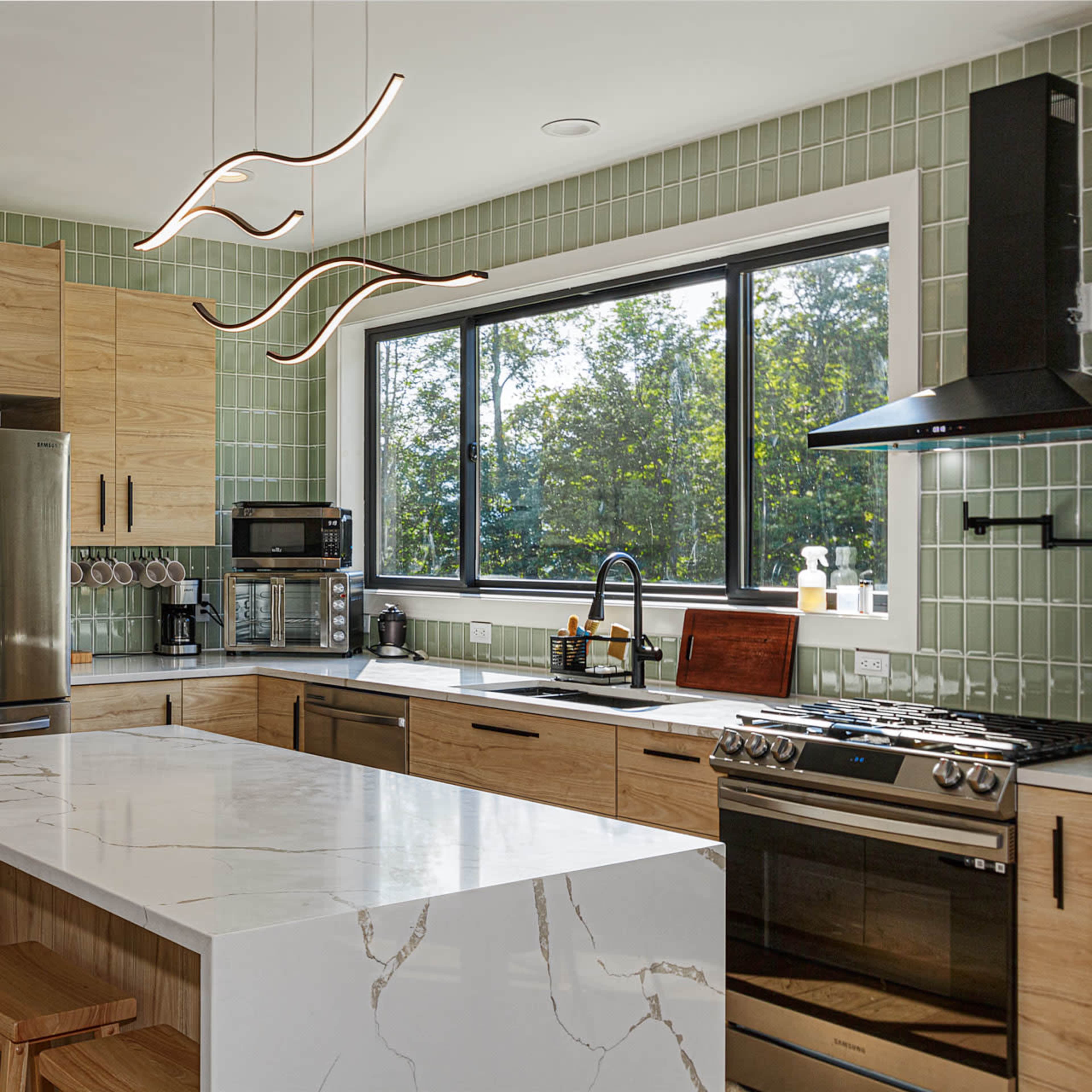 The kitchen features wood cabinetry, stainless steel appliances, a large white marble island, and a window overlooking greenery.