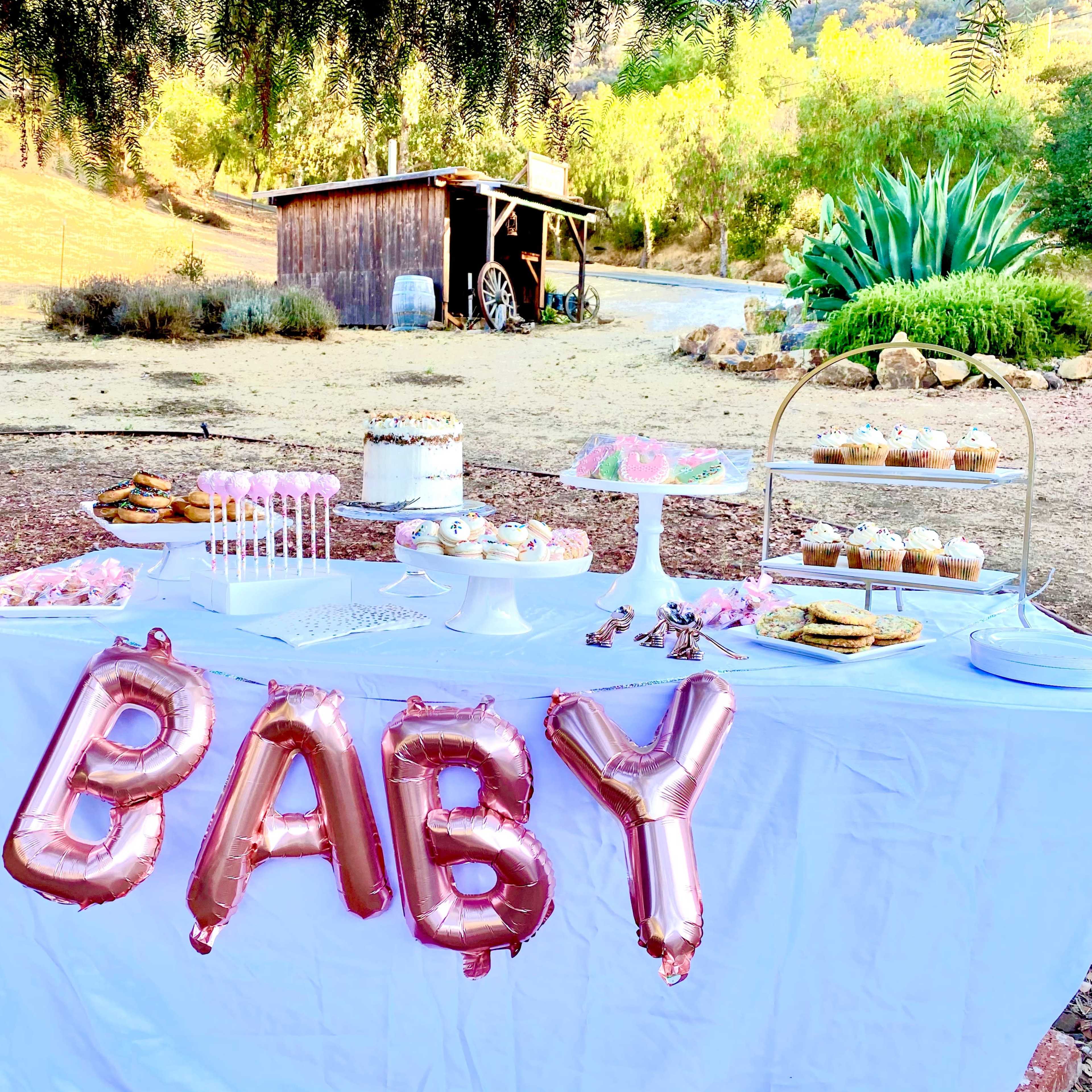 A dessert table featuring a variety of sweets, including a cake and cookies, is decorated with "BABY" balloons in a rural outdoor setting.