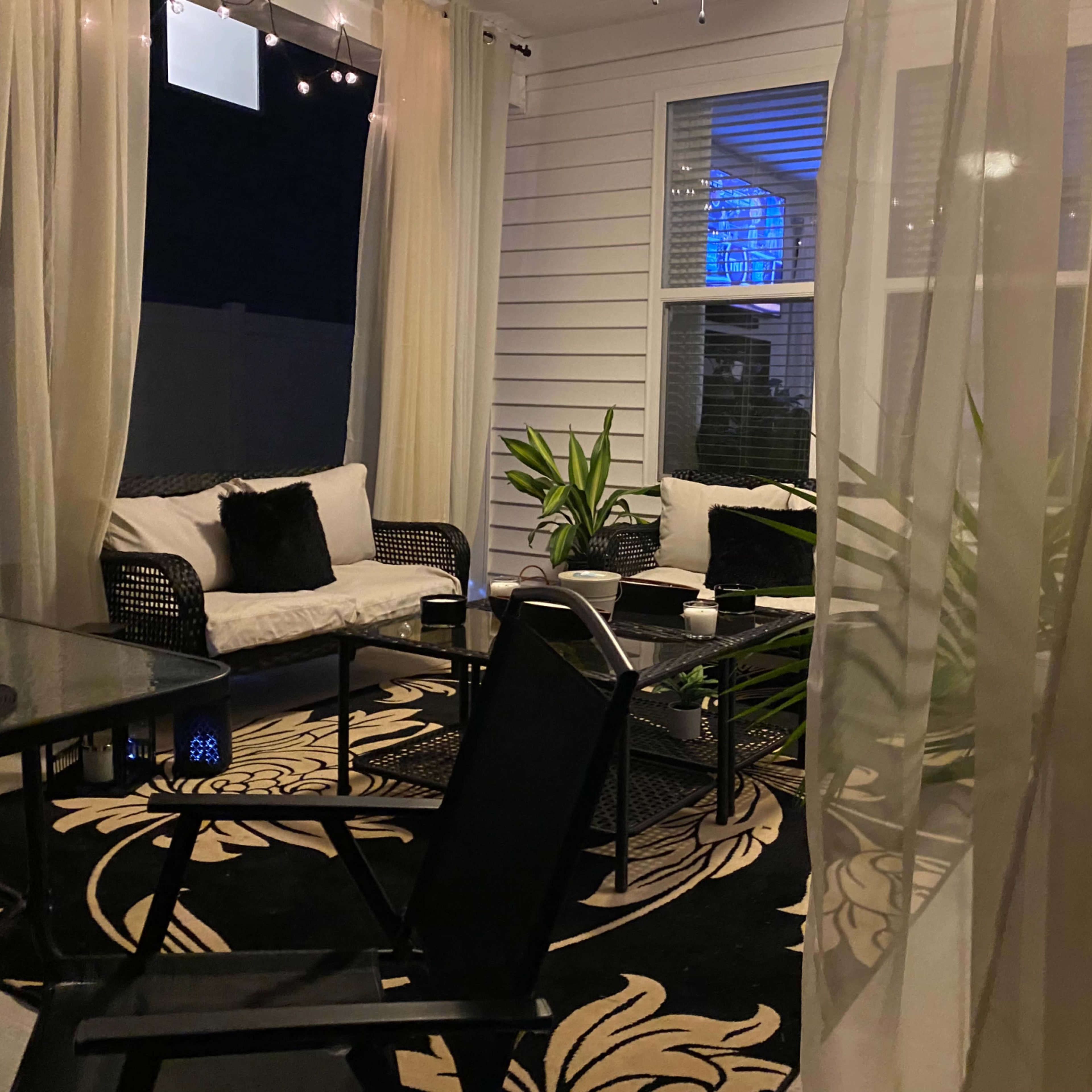 A patio area with black furniture, a patterned rug, and sheer curtains, illuminated softly at night.