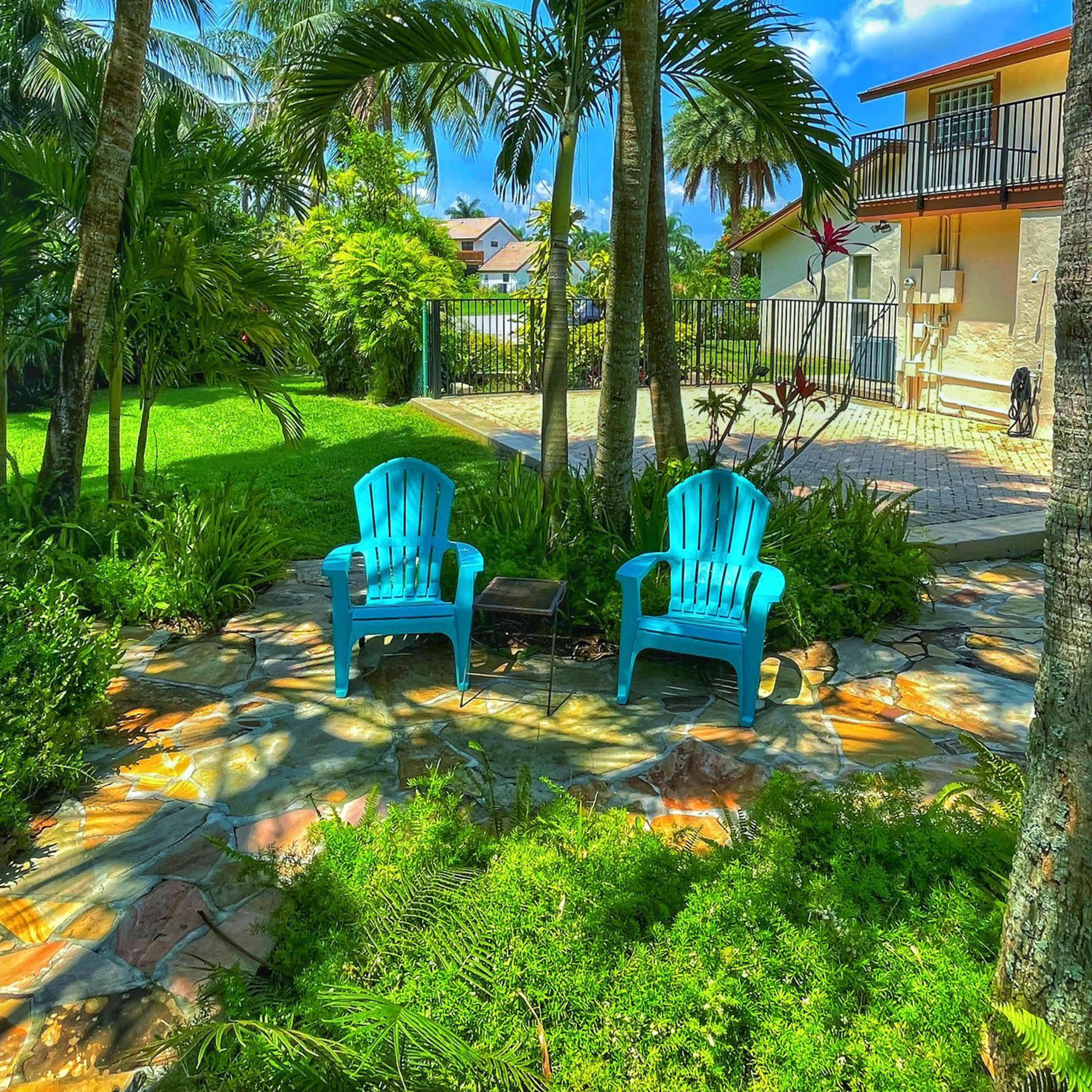 Two blue Adirondack chairs are positioned on a stone patio surrounded by tropical plants and palm trees in a lush garden.