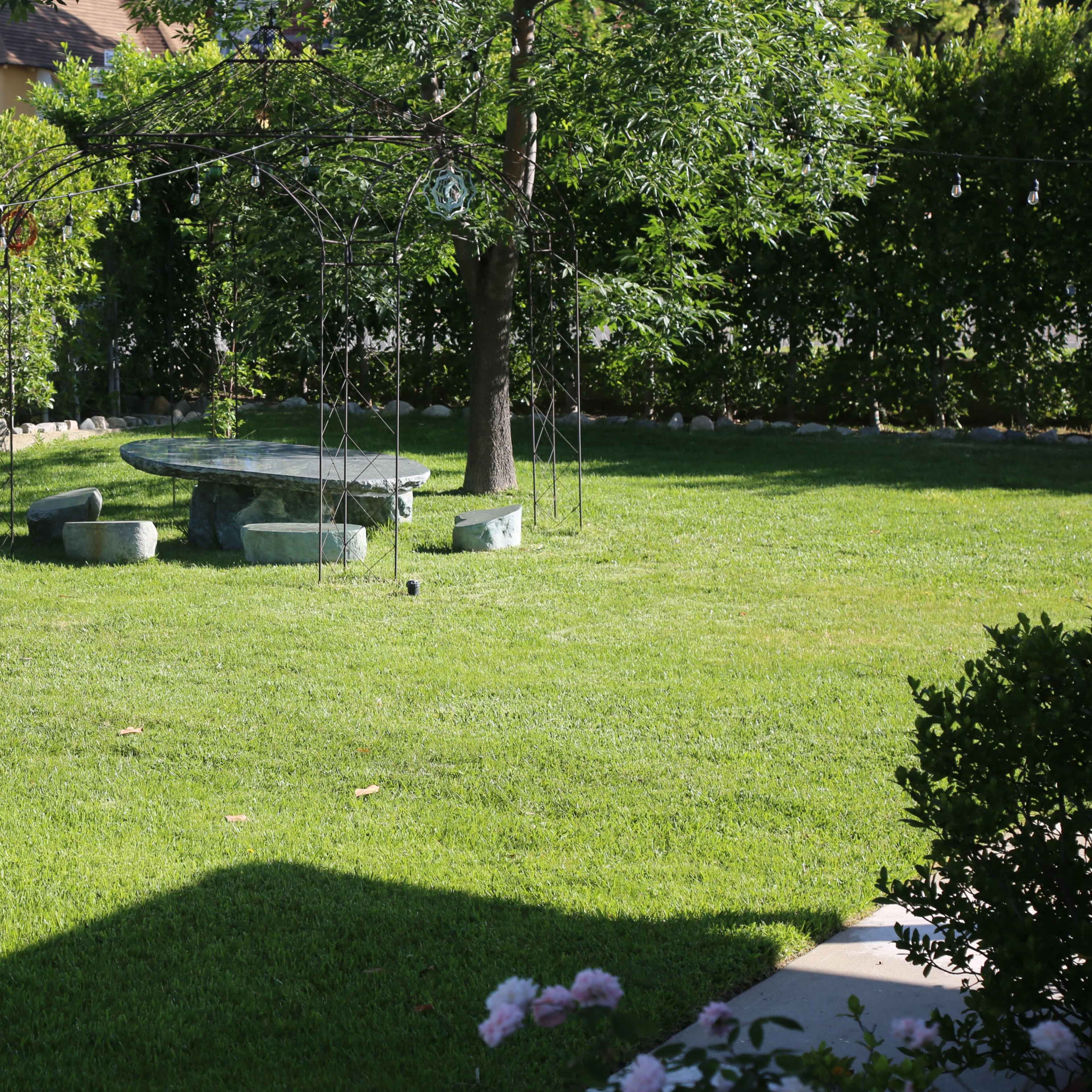 A grassy yard features a stone table with matching benches beneath a tree, surrounded by neatly trimmed hedges.