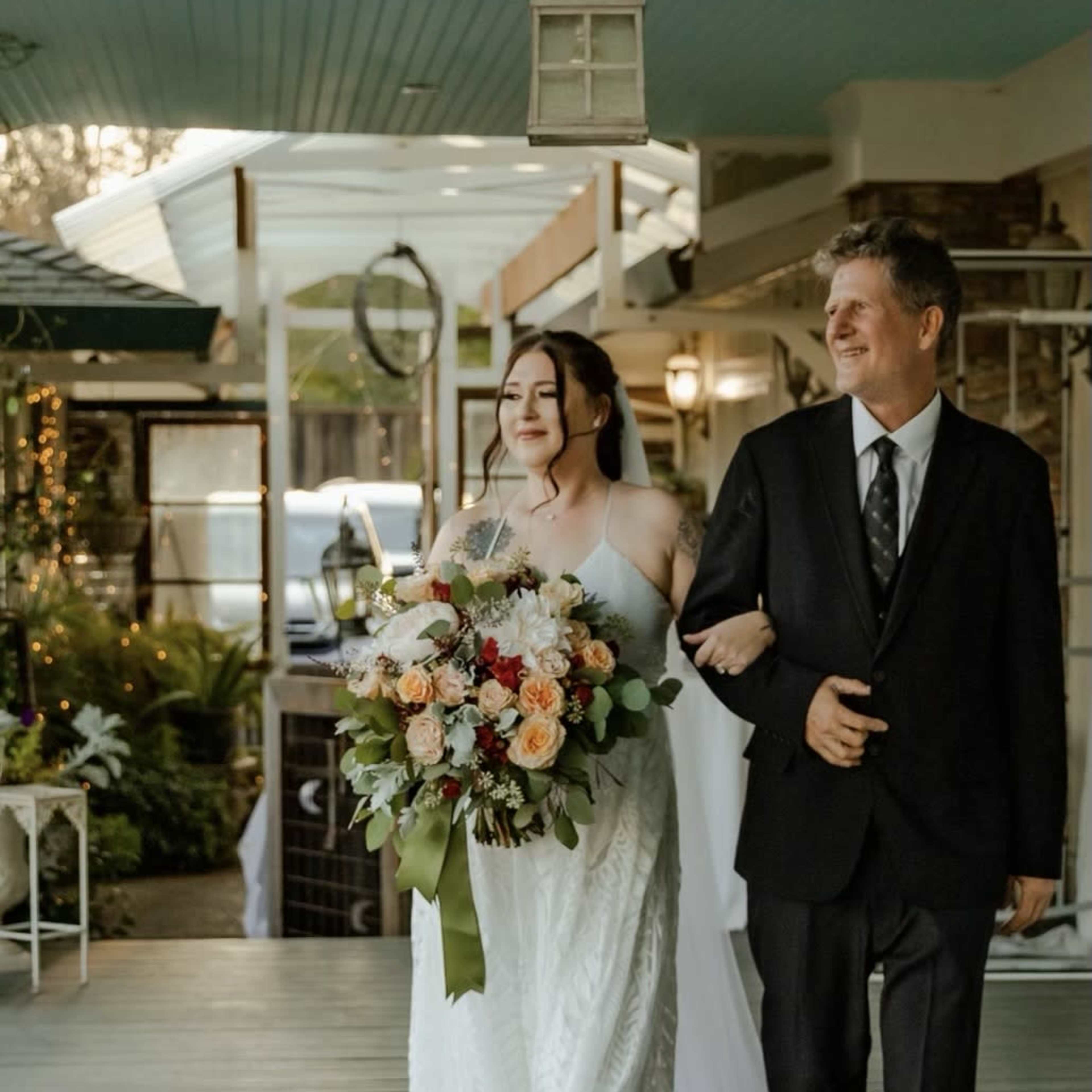 A bride in a white dress walks down a porch holding a bouquet of flowers, accompanied by a man in a black suit.