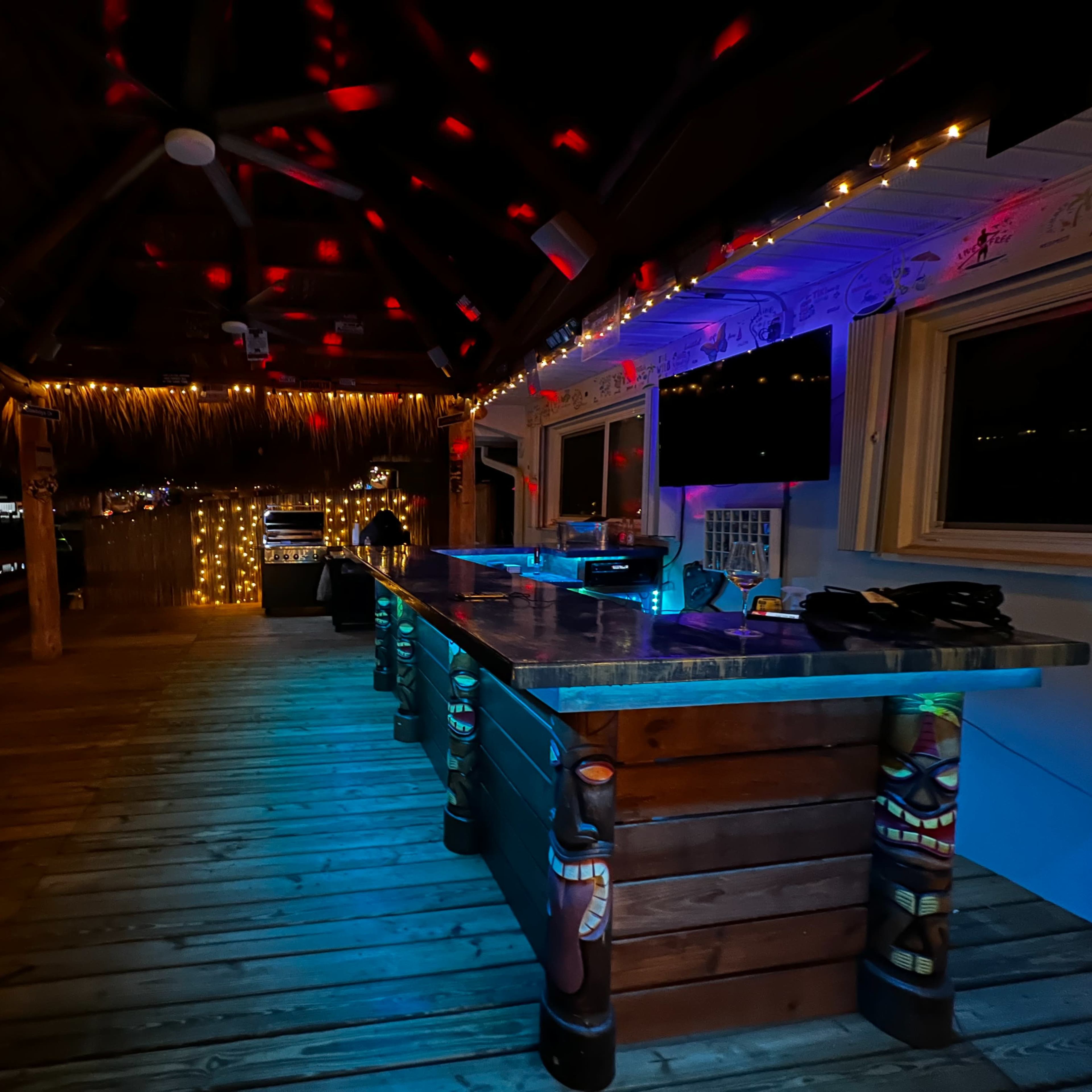 A dimly lit outdoor bar area with wooden furniture, colorful lights, and a thatched roof.
