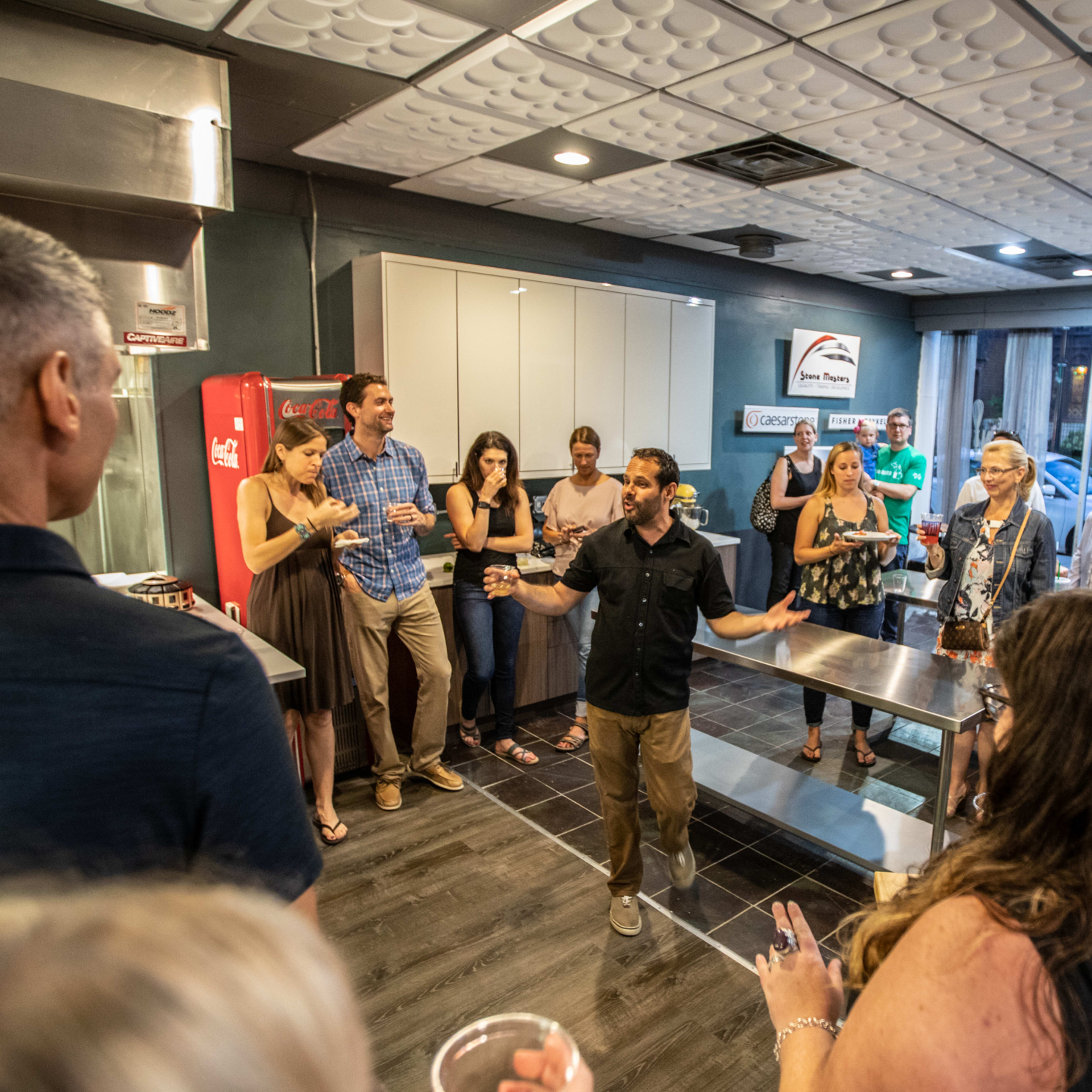 A group of people gathers in a modern kitchen space, with some standing around a central island while a man speaks in front of them.