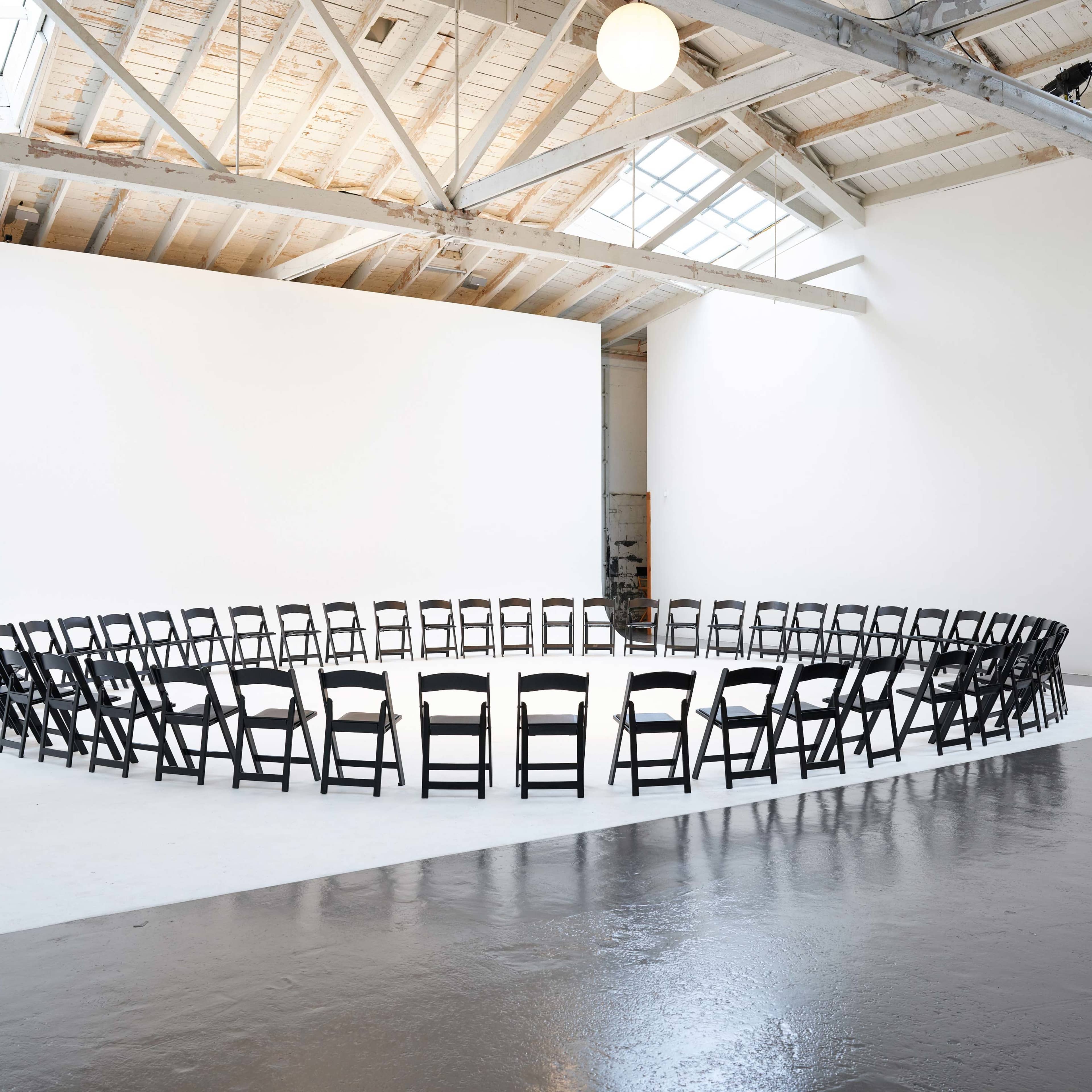 A circle of black chairs is arranged in an empty, well-lit space with white walls and a skylight.